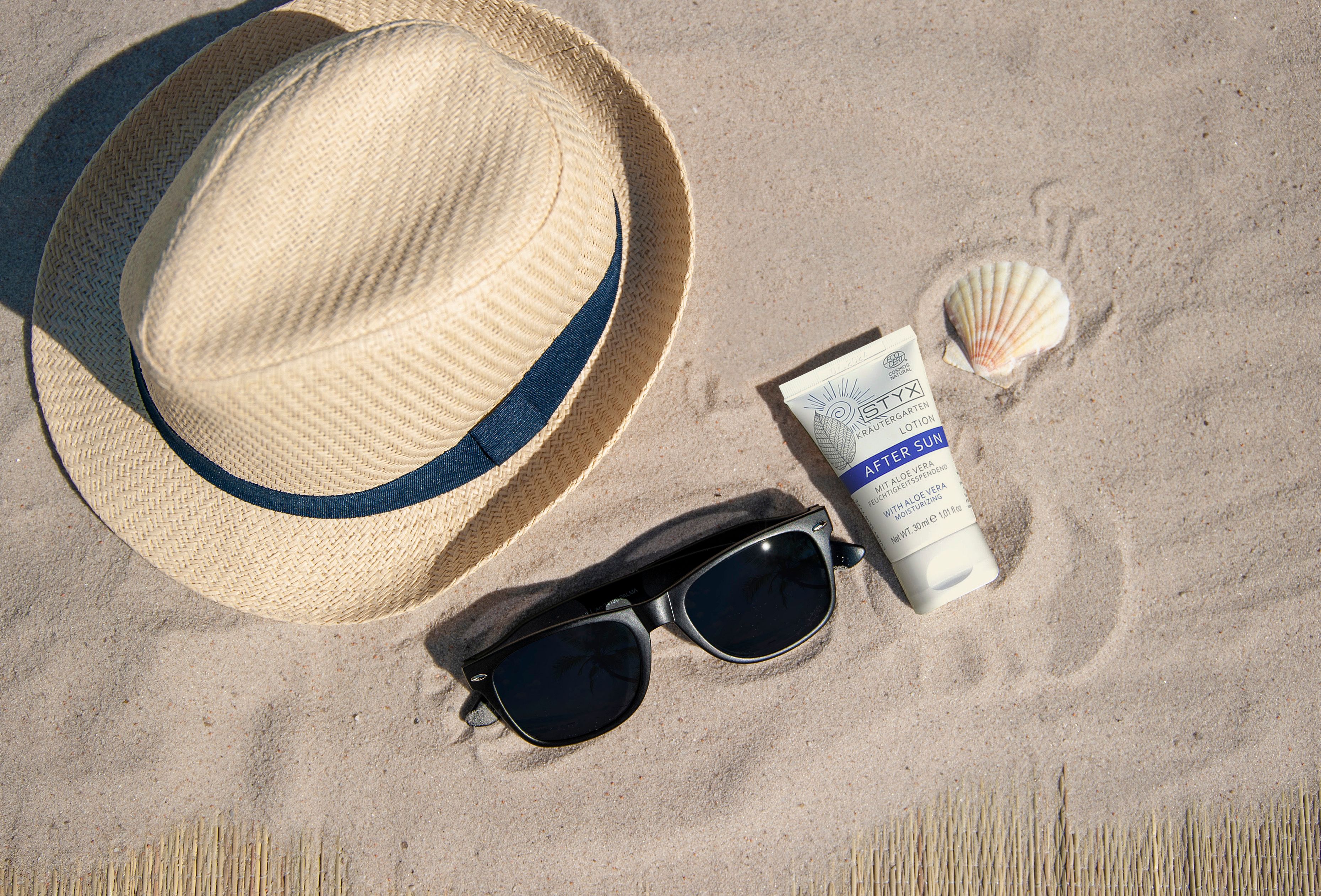 Sonnenhut, Sonnenbrille, Muschel und Tube "AFTER SUN" auf Sand. Tube mit "STYX" und "AFTER SUN" Beschriftung.