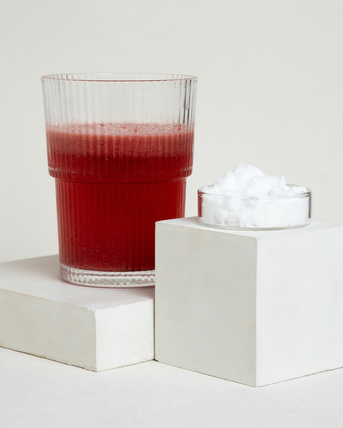 Ein Glas mit rotem Getränk und eine kleine Schale mit weißem Pulver auf weißen Würfeln.