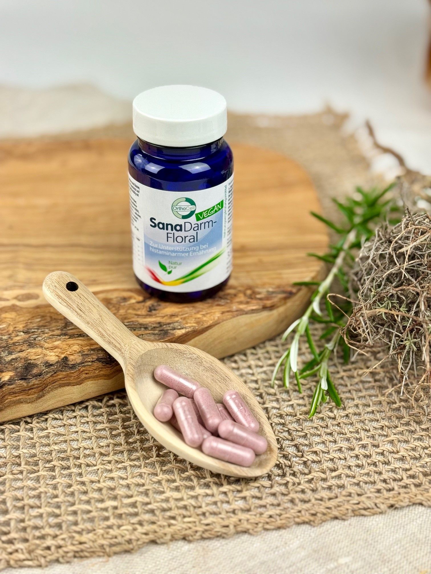 Blaue Flasche SanaDarm-Floral neben Holzlöffel mit Kapseln. Auf Holzbrett und Leinenstoff. Vegan-Siegel.