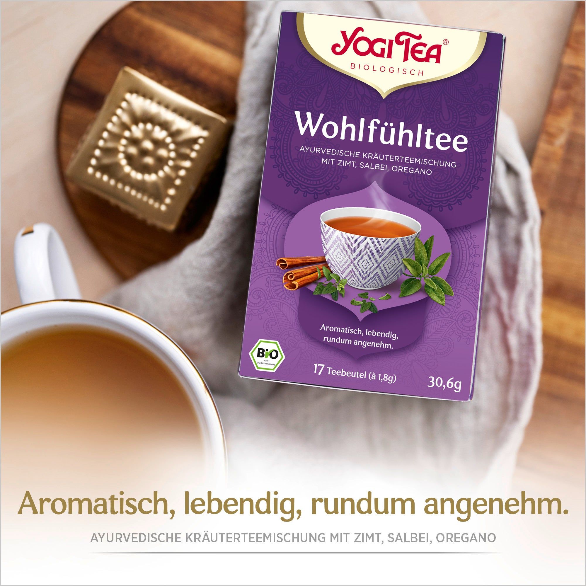 Yogi Tea Wohlfühltee Bio Kräutertee. Lila Verpackung mit Logo, Tasse, Zimtstangen und Kräutern. Auf Holzuntergrund.