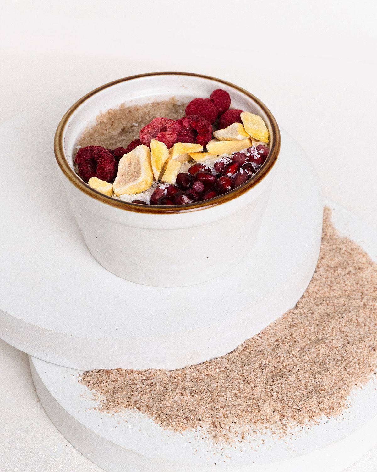Schale mit Müsli, Himbeeren, Granatapfelkernen und Ananasstücken. Auf weißem Untergrund. Daneben Flohsamenschalen.