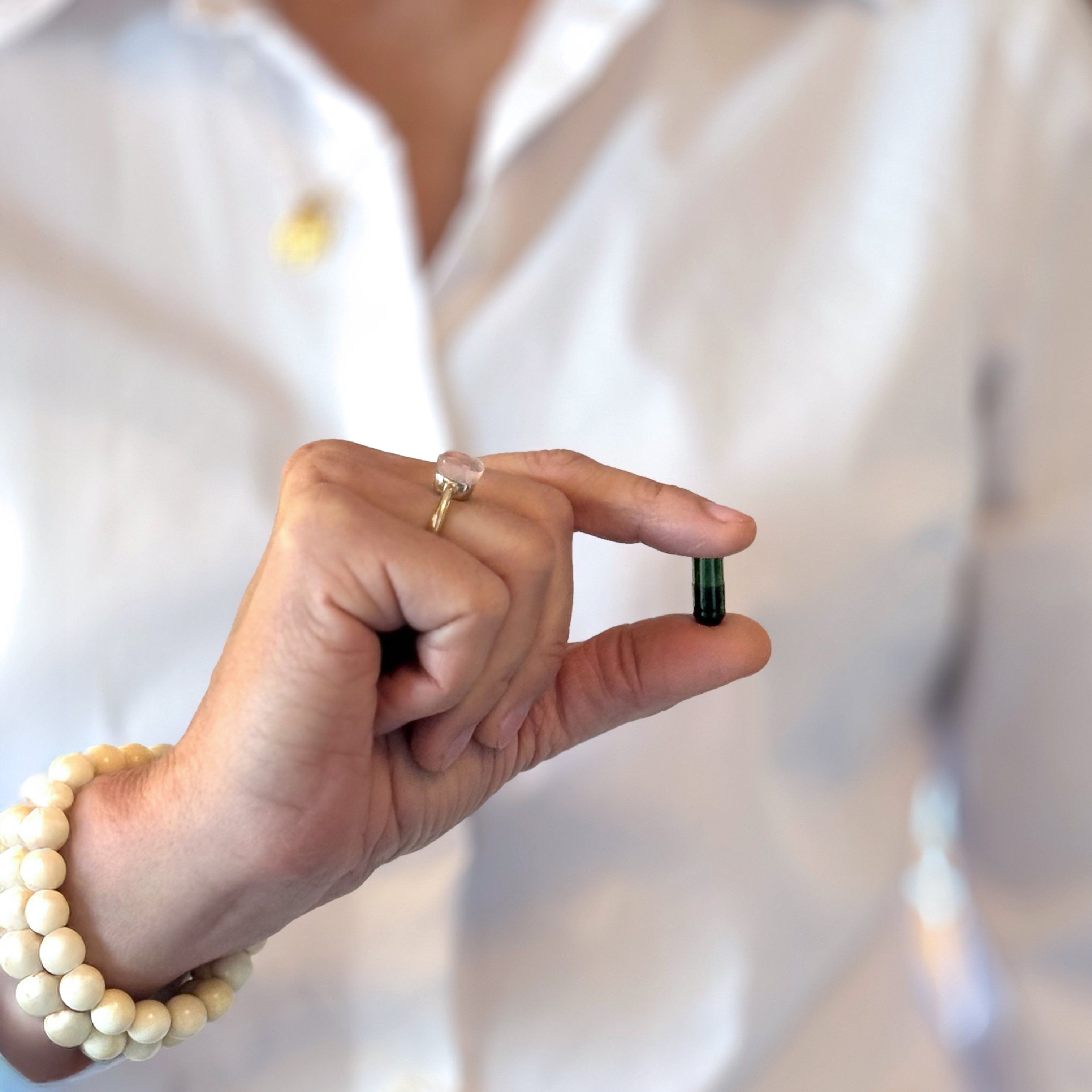 Hand hält eine grüne Kapsel. Person trägt Schmuck.