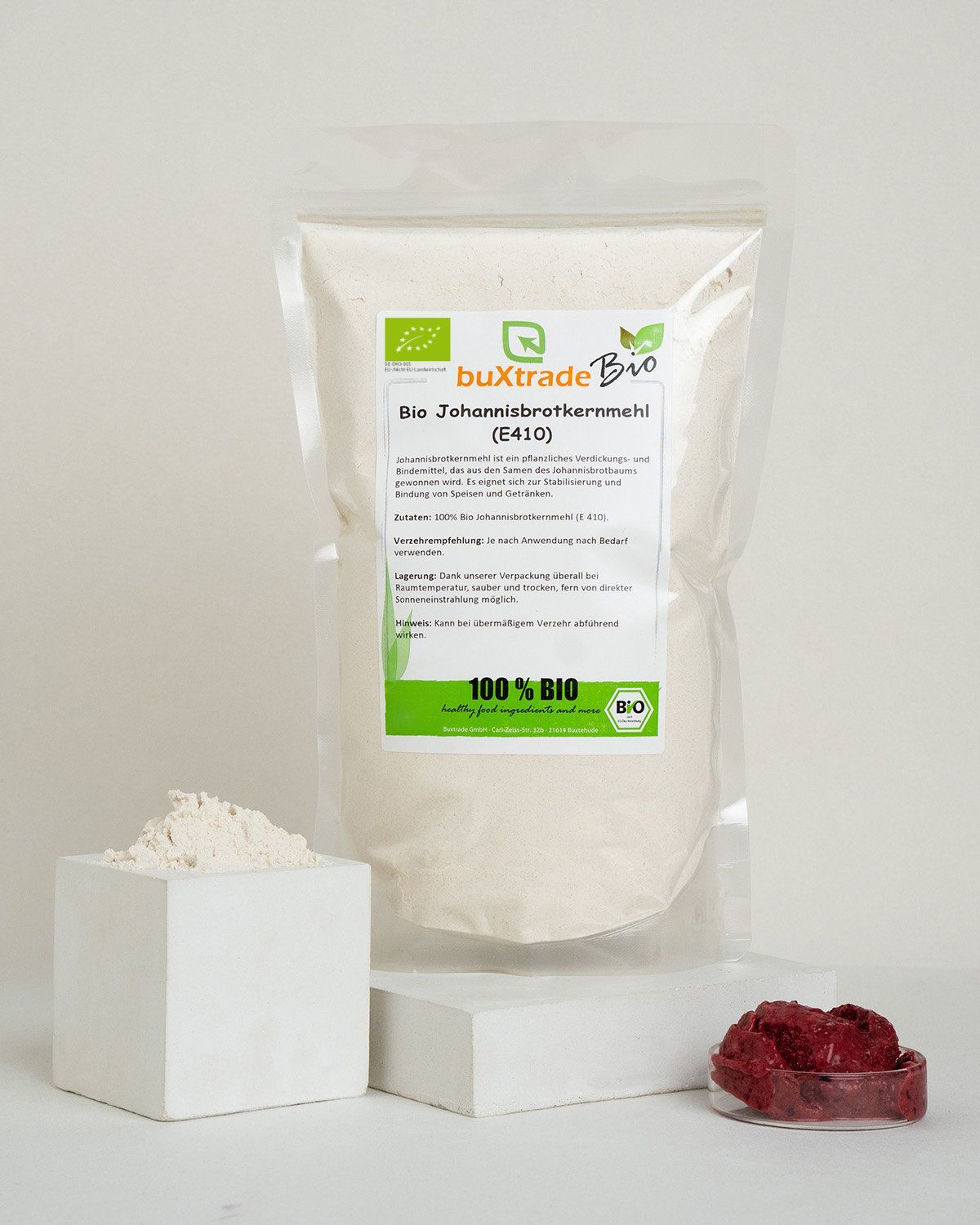 Bio Johannisbrotkernmehl in einem Beutel und einem weißen Würfel, daneben rote Beeren.
