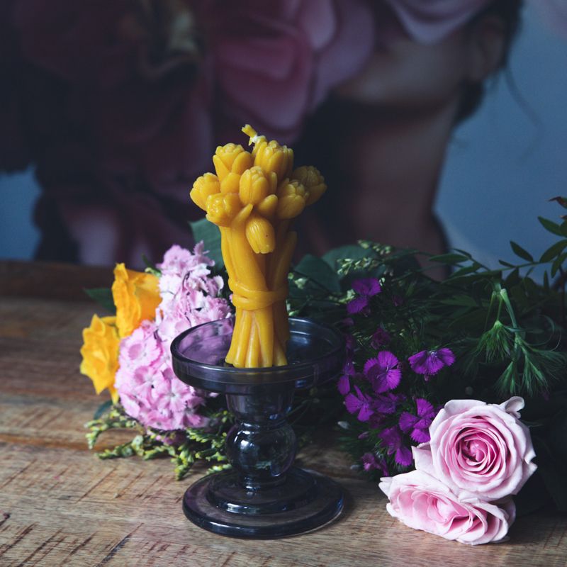 Brennende gelbe Wachskerze in Tulpenform auf einem Kerzenständer, umgeben von Blumen.