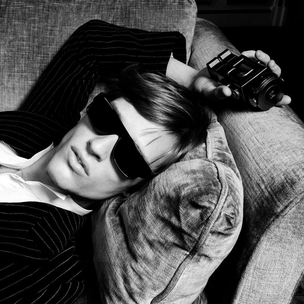 Schwarz-Weiß-Foto eines Mannes in Anzug und Sonnenbrille, der sich auf einem Sofa ausruht, mit einem Parfümflakon.