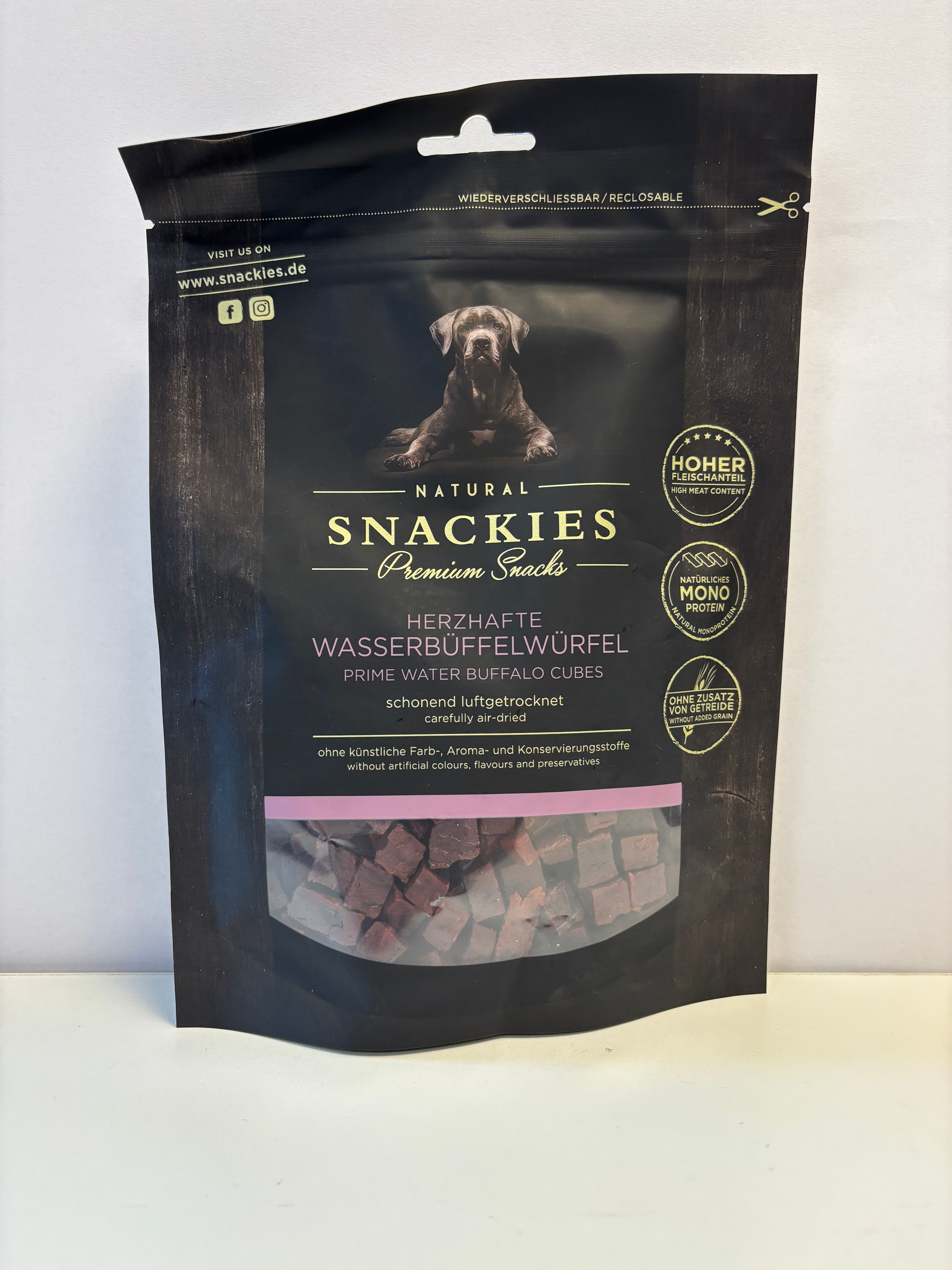 Verpackung mit Snackies Herzhafte Wasserbüffelwürfel. Schwarze Tüte mit Produktbild, Text und Siegeln. Sichtfenster zeigt Würfel.