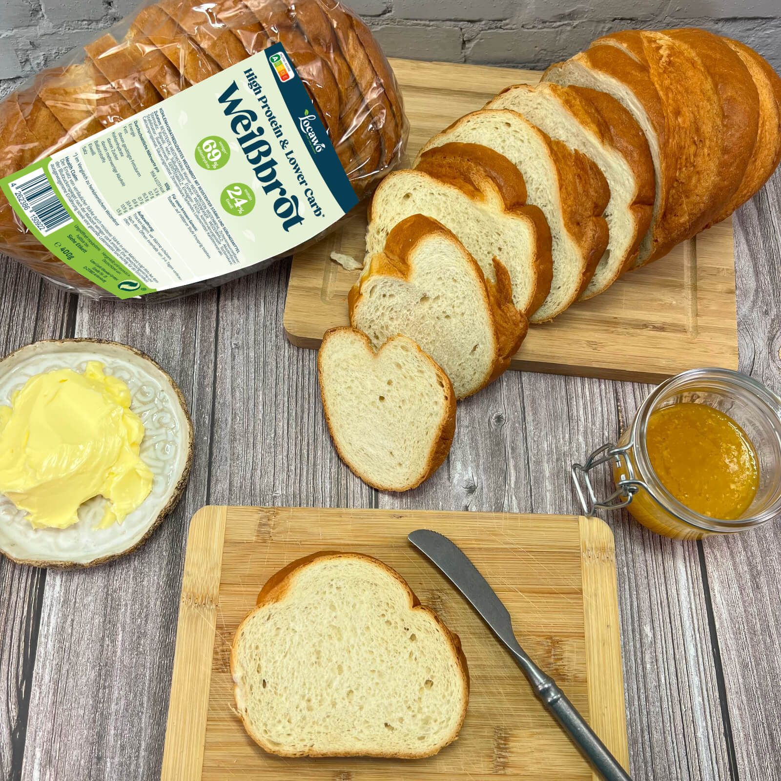 Weißbrotscheiben, Butter, Marmelade und Messer auf Holzbrettern. Im Hintergrund: verpacktes Brot. Aufschrift: Locawo Weißbrot.