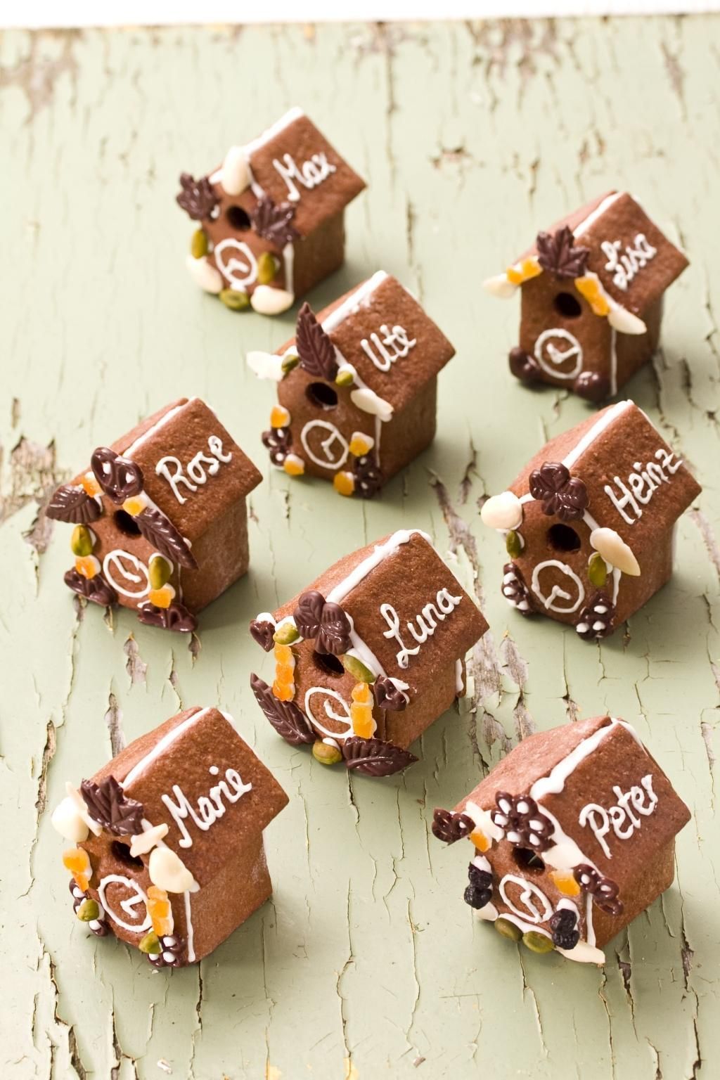 Acht Lebkuchenhäuser mit Namen. Dekorationen: Nüsse, Blätter, Zuckerguss. Auf grünem Untergrund.