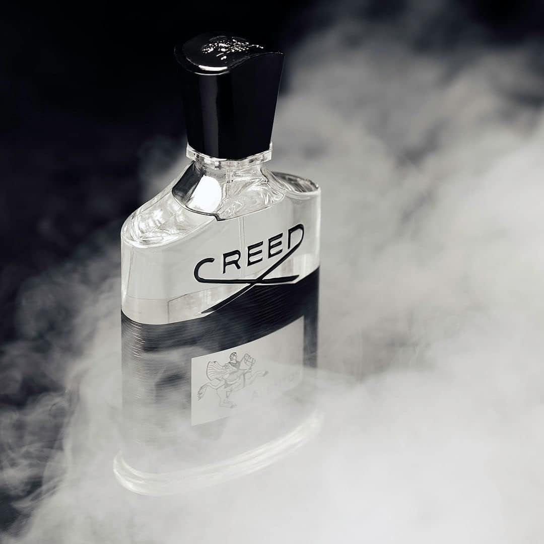 Glasflakon mit schwarzem Verschluss. Aufschrift Creed. Etikett mit AVENTUS. Nebeliger Hintergrund.