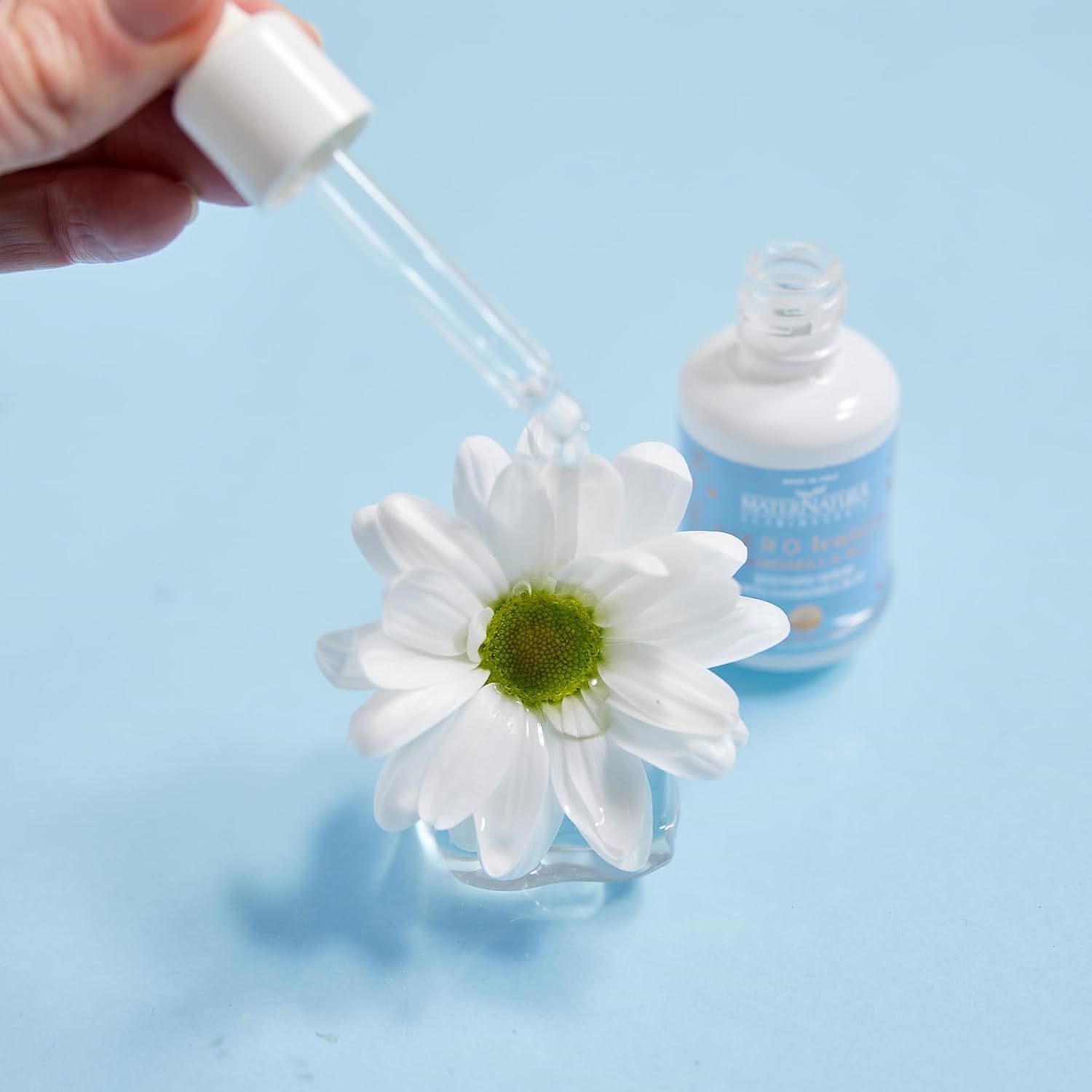 Eine Pipette gibt einen Tropfen Serum auf eine weiße Blüte. Ein kleiner Flakon steht dahinter.
