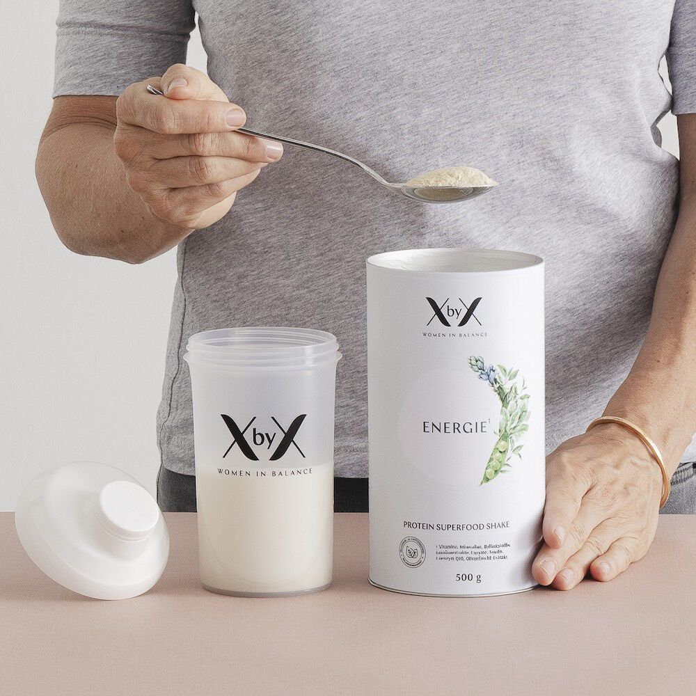 Person hält Löffel mit Pulver über Shaker und Dose mit 'XbyX Energie'.