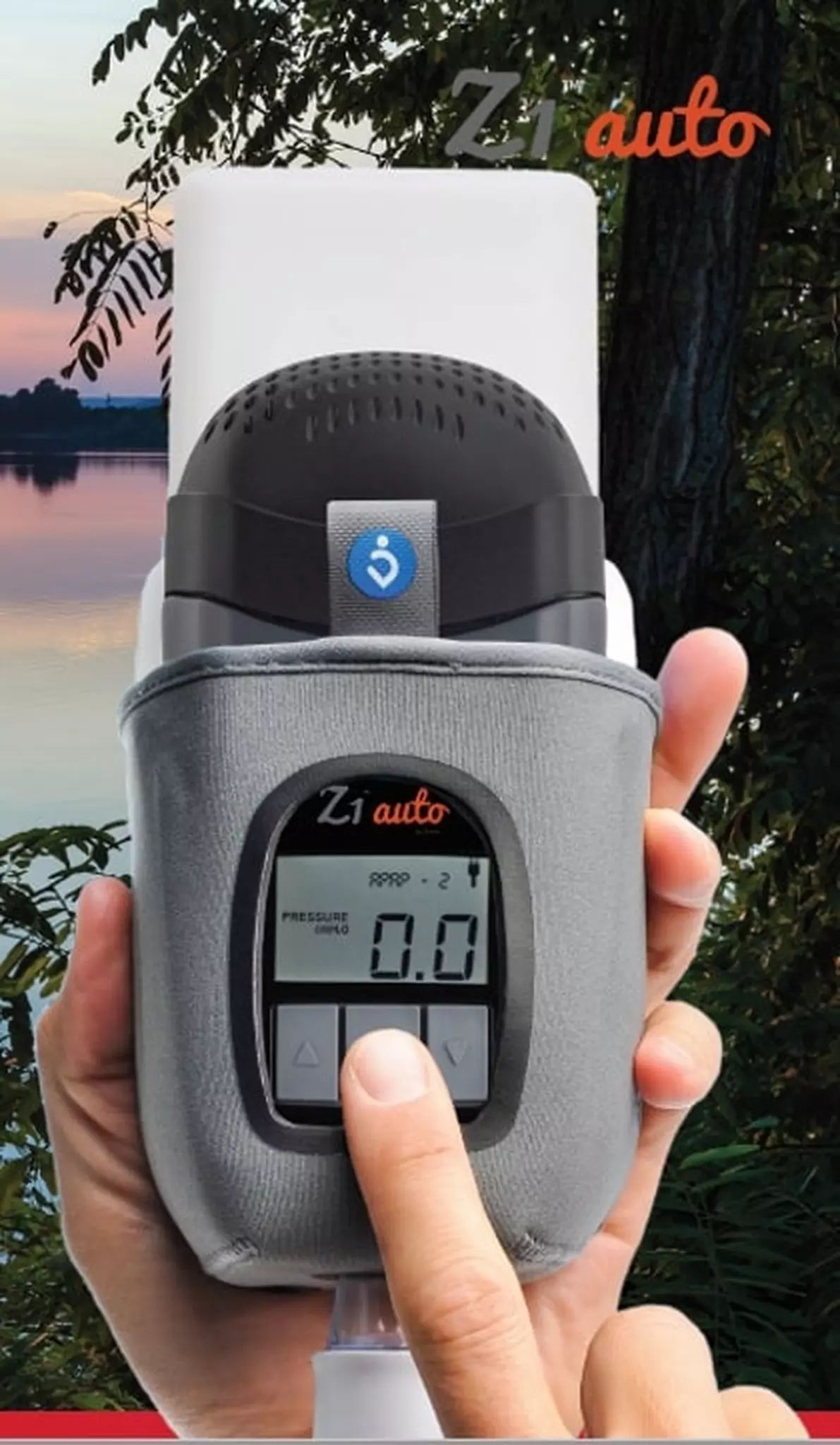 Graues Reise-CPAP-Gerät mit Display, von Händen gehalten, vor See und Bäumen.