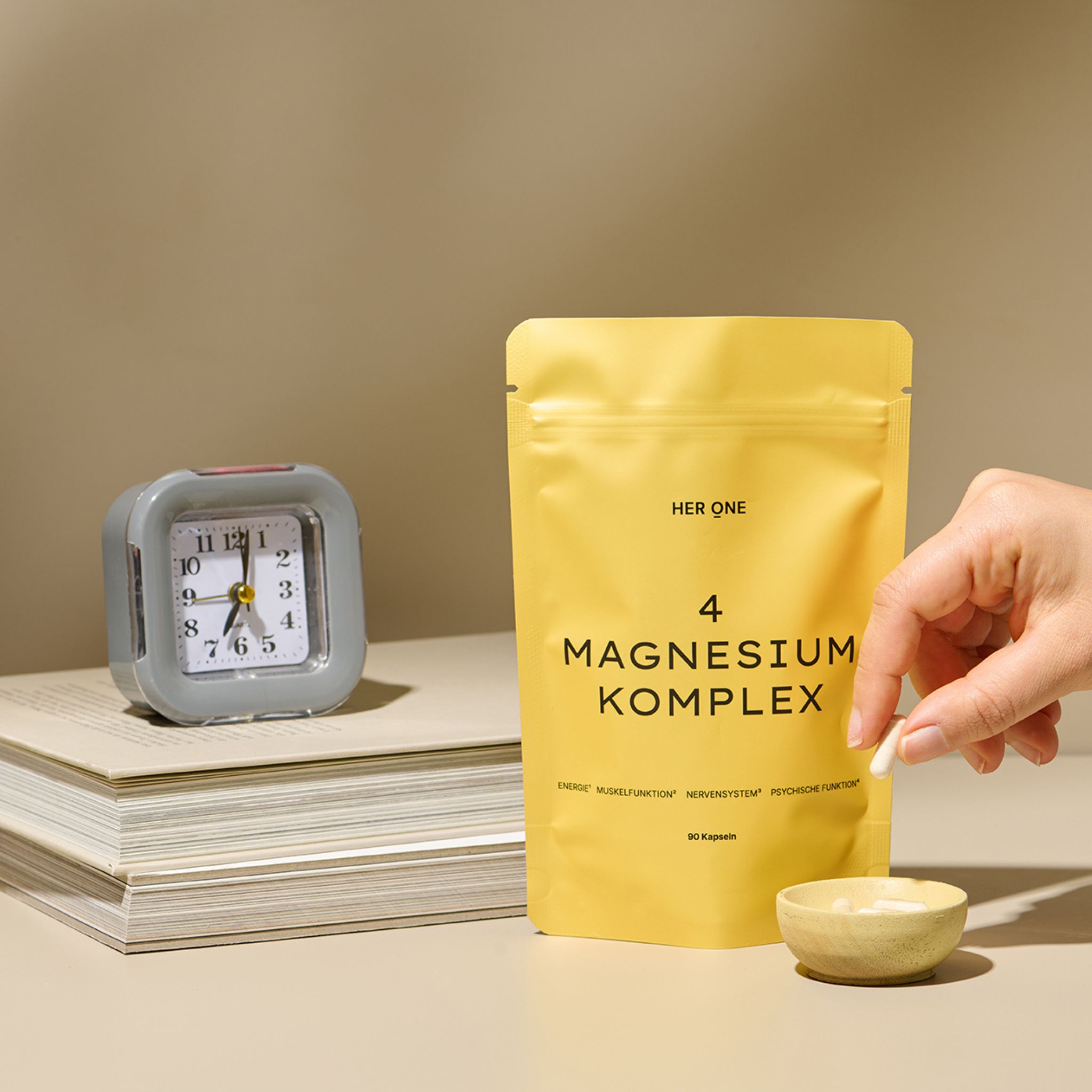 Gelbe Verpackung HER ONE 4 MAGNESIUM KOMPLEX neben Uhr und Büchern. Hand hält Kapsel über kleiner Schale.