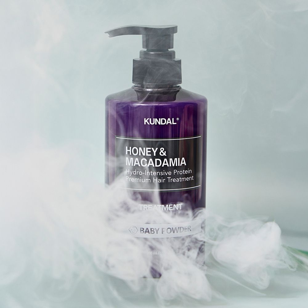 Violette Flasche mit schwarzem Pumpverschluss, umgeben von weißem Rauch. Auf dem Etikett steht "KUNDAL Honey & Macadamia Baby Powder Treatment".