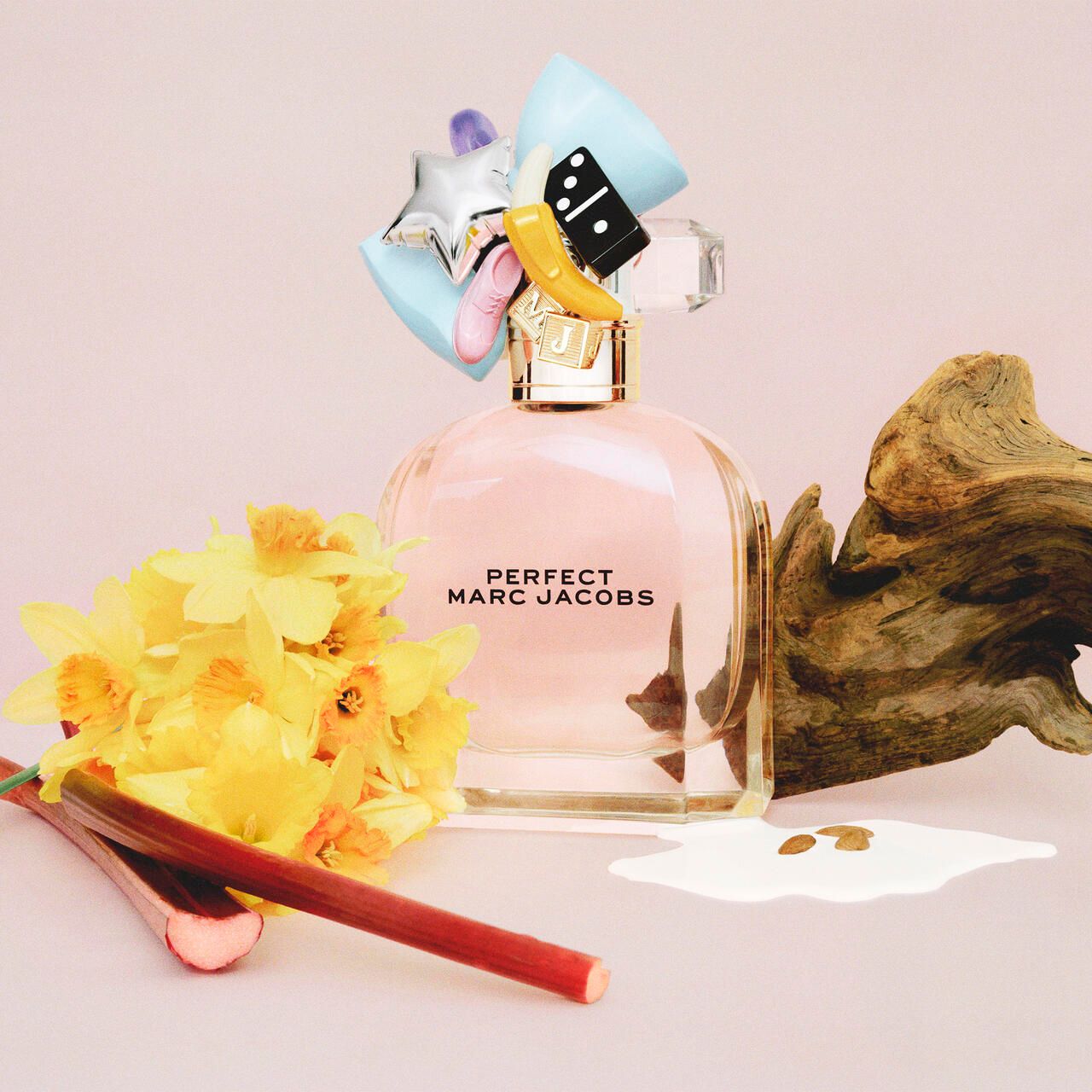 Parfümflakon mit dekorativem Deckel. Aufschrift: PERFECT MARC JACOBS. Neben dem Flakon: Blumen, Holzstück, Flüssigkeit.