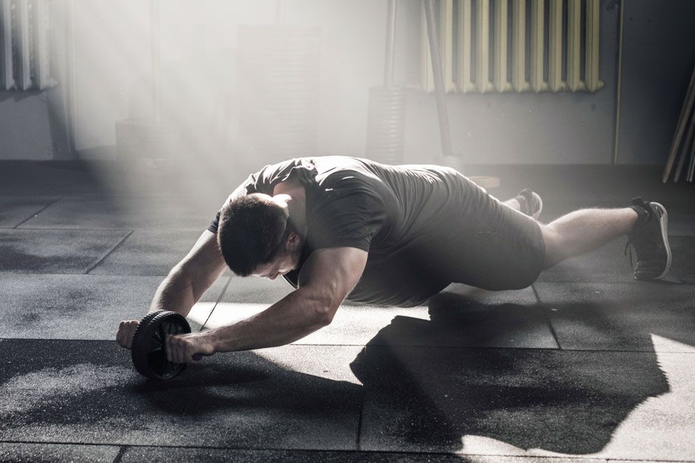 Mann trainiert mit Fitnessroller. Schwarzes Rad, Griffe. In Sporthalle. Lichtstrahlen. Auf dem Boden liegend.
