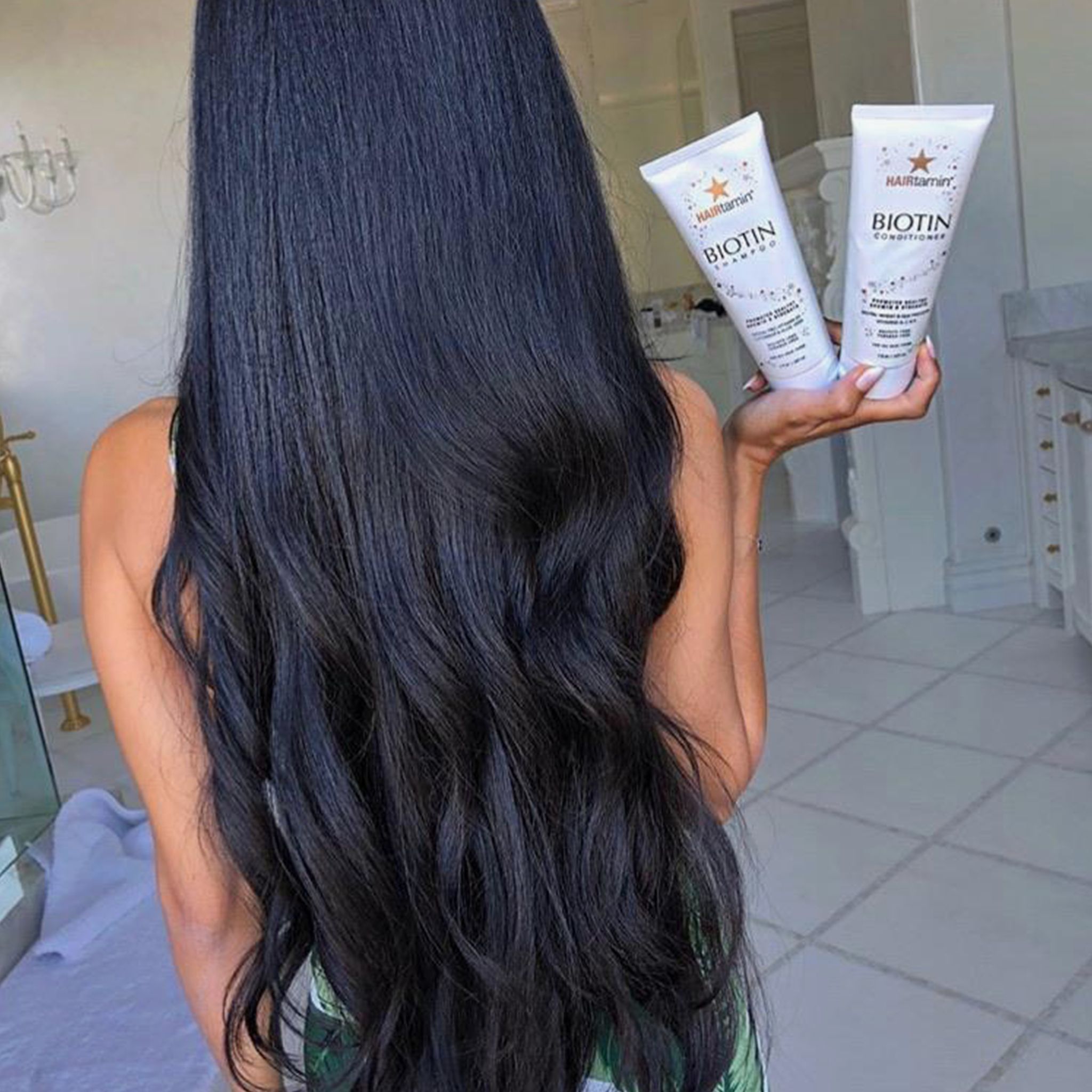 Person hält zwei Tuben "HAIRtamin Biotin Conditioner". Lange, schwarze Haare im Vordergrund. Badezimmerhintergrund.