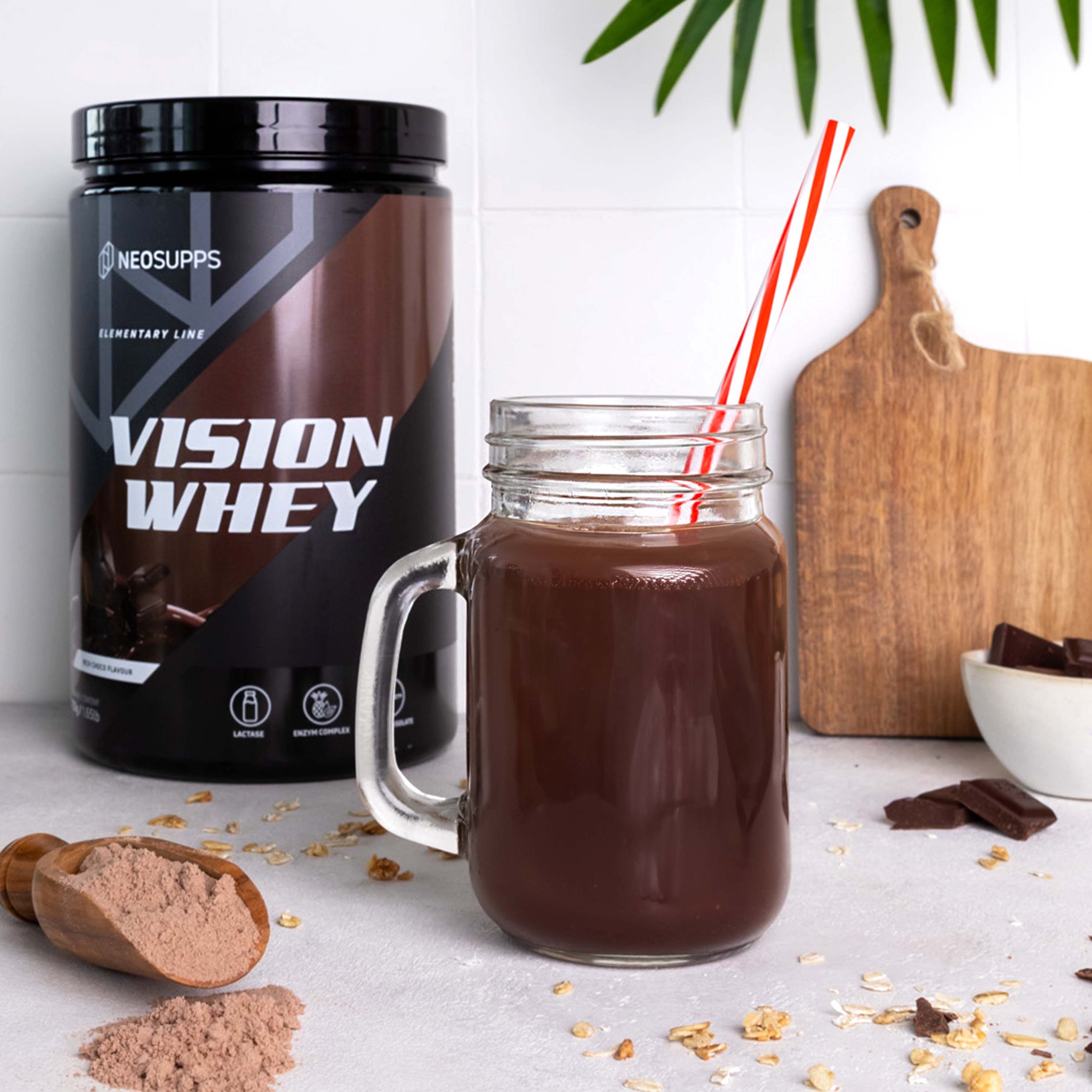 Glas mit Schokoladengetränk, Dose Vision Whey daneben. Aufschrift: Vision Whey. Strohhalm im Glas. Löffel mit Pulver daneben.