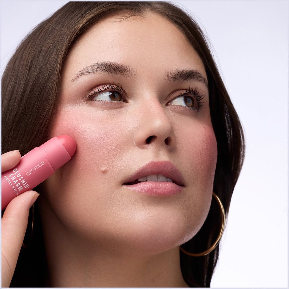 Frau trägt Catrice Blushin' Charm Multi Stick auf die Wange auf. Rosa Stick, Produktname und Marke sichtbar.