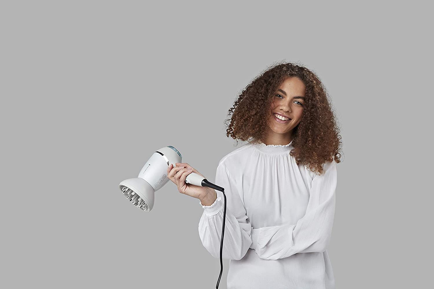 Frau hält weißen Haartrockner. Grauer Hintergrund. Sie trägt ein weißes Oberteil.