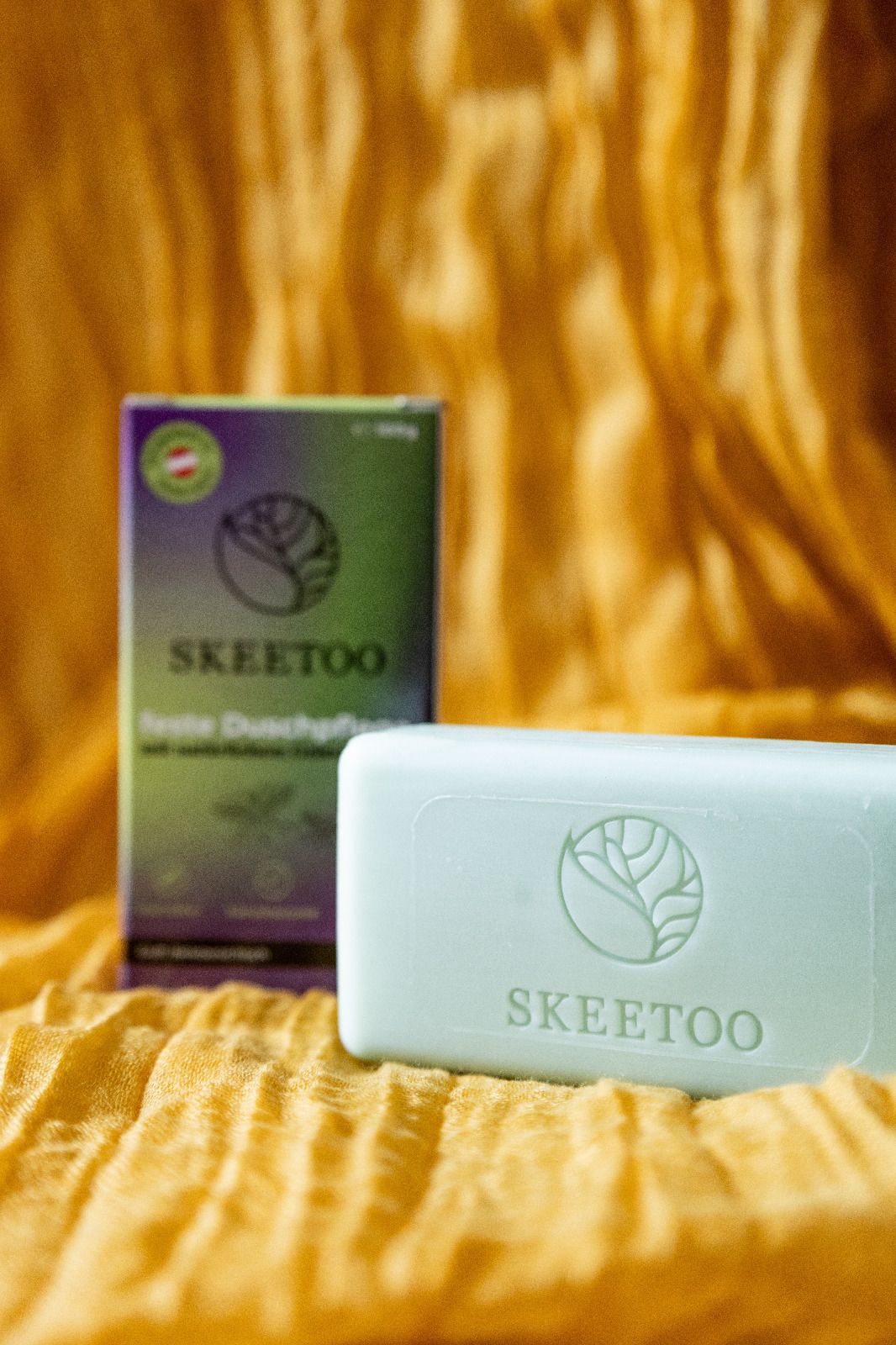 Hellgrünes Seifenstück und Verpackung mit Logo und Schriftzug SKEETOO. Verpackung mit Aufdruck. Seife liegt auf gelbem Stoff.