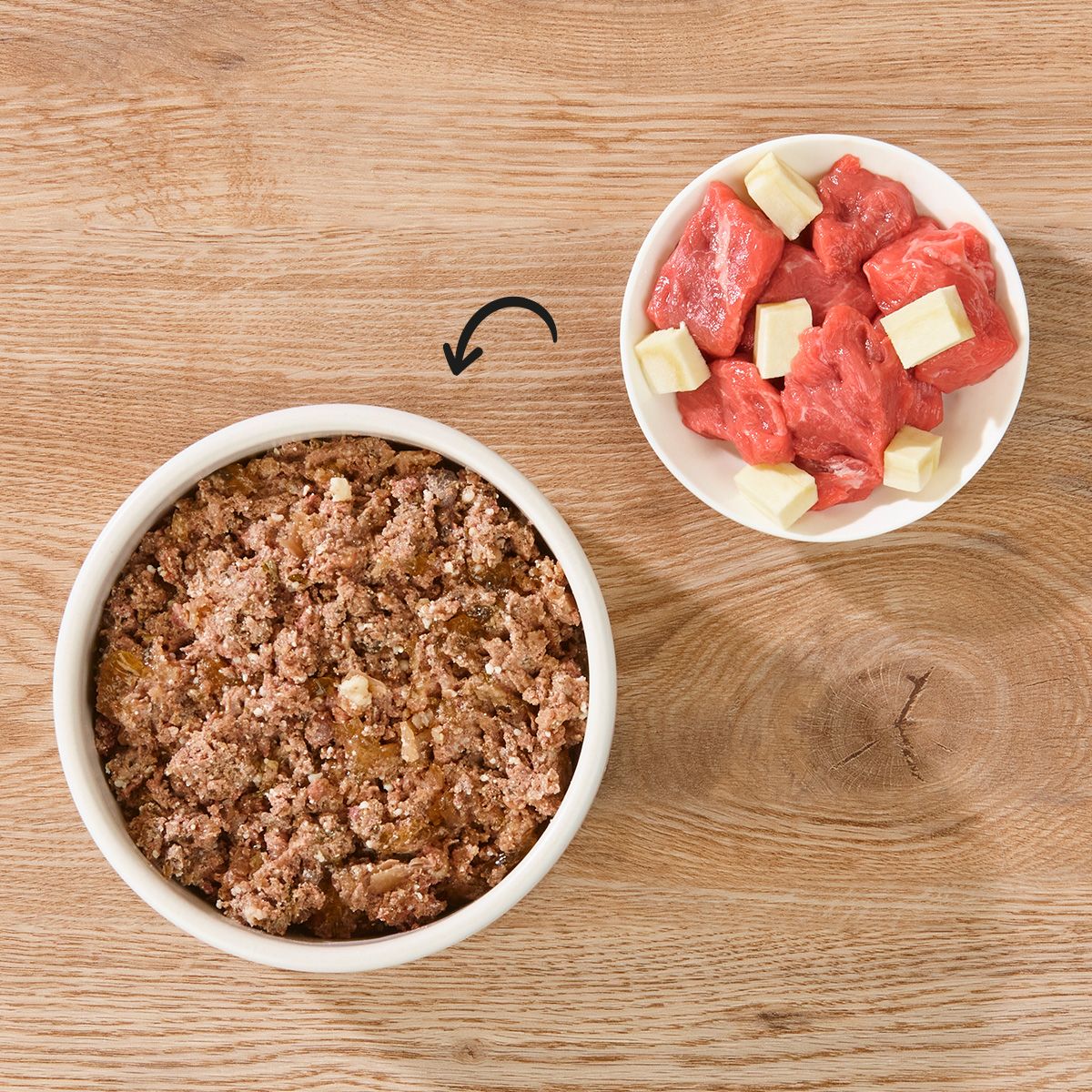 Zwei Schalen auf Holz. Eine mit Futter, eine mit Fleisch und Käsewürfeln. Pfeil zeigt von Fleisch zu Futter.