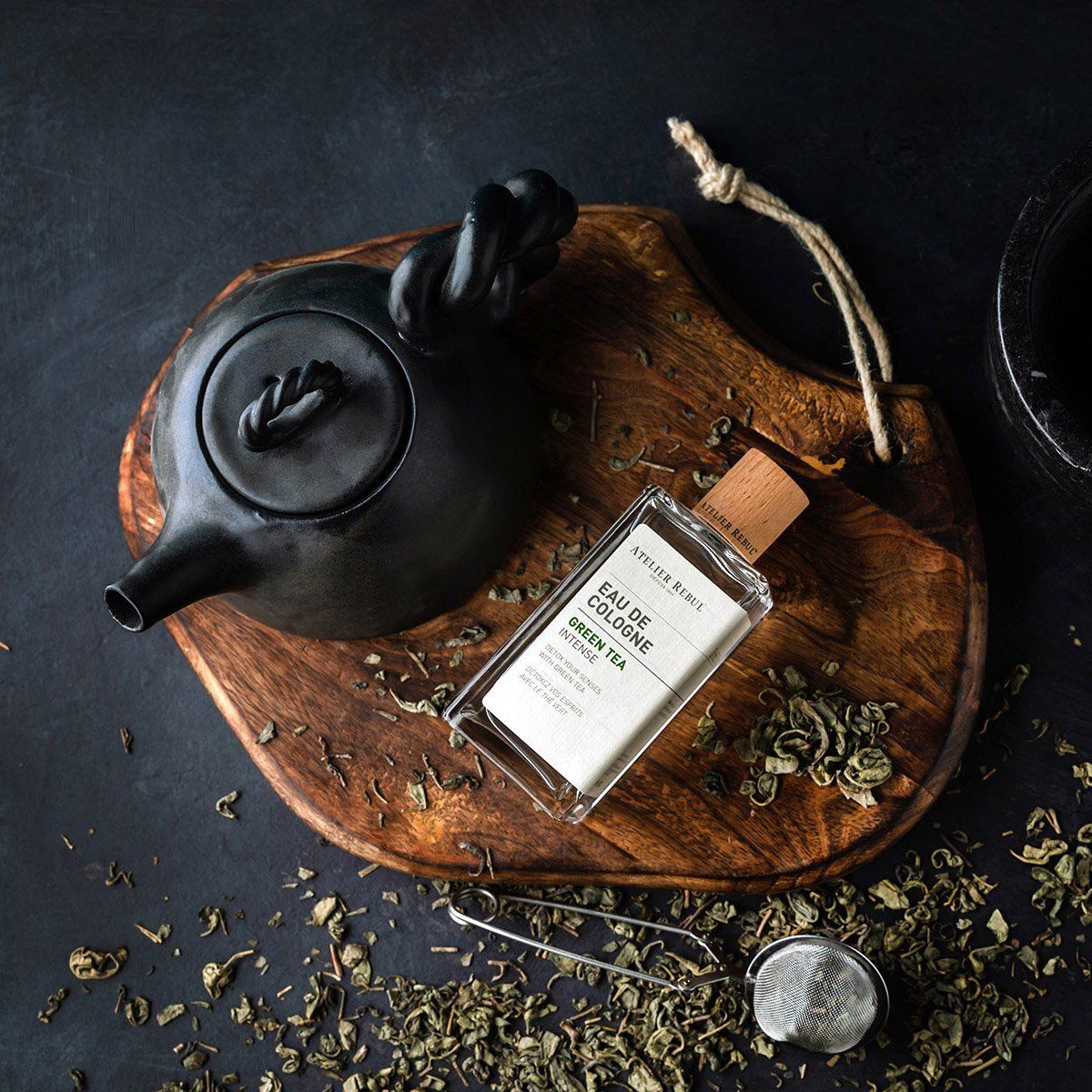 Glasflakon mit Holzverschluss auf Holzbrett. Aufschrift: Atelier Rebul Eau de Cologne Green Tea Intense. Neben schwarzer Teekanne und Teesieb.