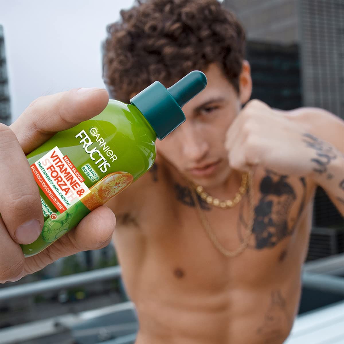 Person hält grüne Flasche mit Tropfer. Aufschrift: Garnier Fructis Vitamine & Kraft. Mann mit Tattoos und Halskette im Hintergrund.