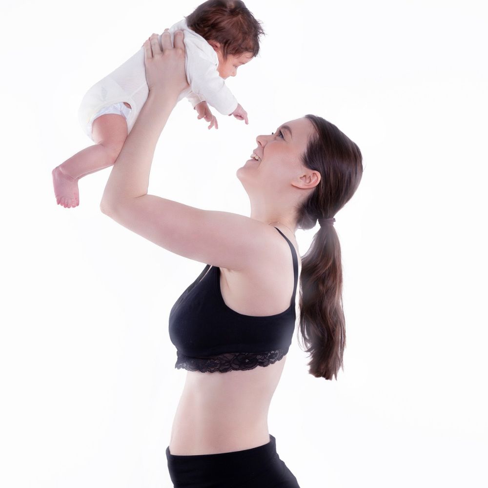 Frau in schwarzem Still-BH hebt Baby hoch. Schwarze Hose. Weißer Hintergrund. Frau lächelt.