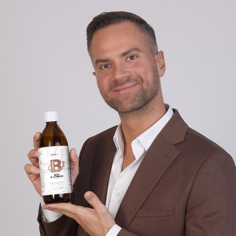 Mann hält braune Flasche "BEST LAB B-COMPLEX". Er trägt einen braunen Anzug. Helles Hintergrund.
