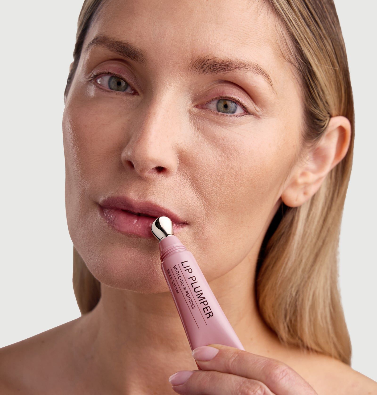 Frau trägt Lip Plumper auf. Tube mit Aufschrift: LIP PLUMPER WITH CHILI & PEPTIDES. Applikator am Mund.