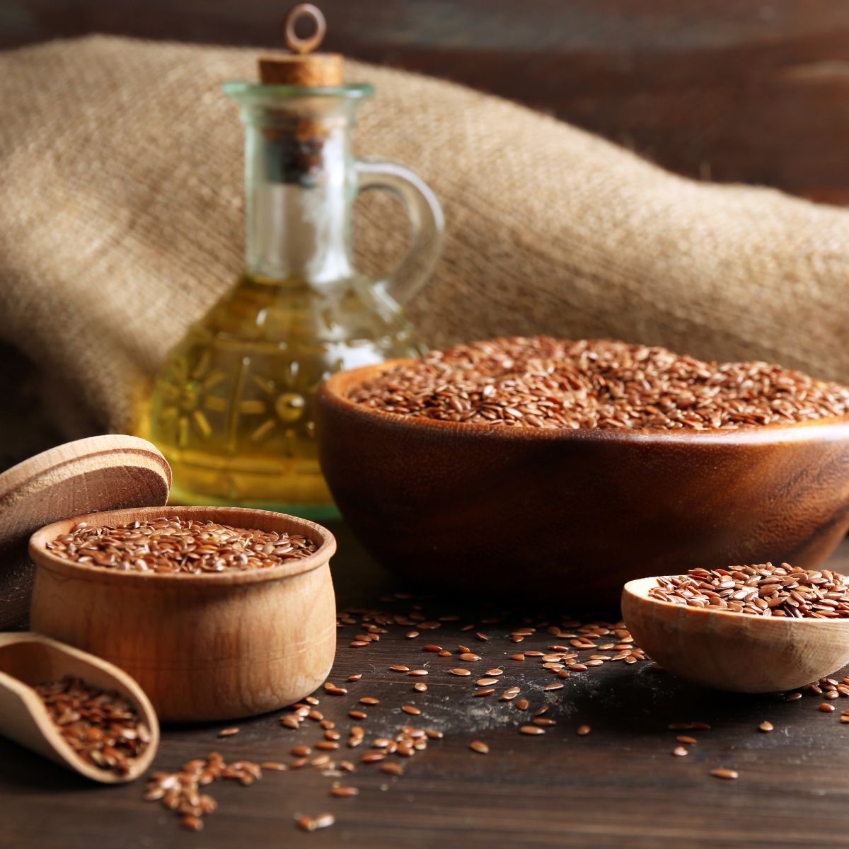 Holzschalen mit braunen Leinsamen, daneben Ölflasche und Löffel. Hintergrund: grobes Leinentuch.