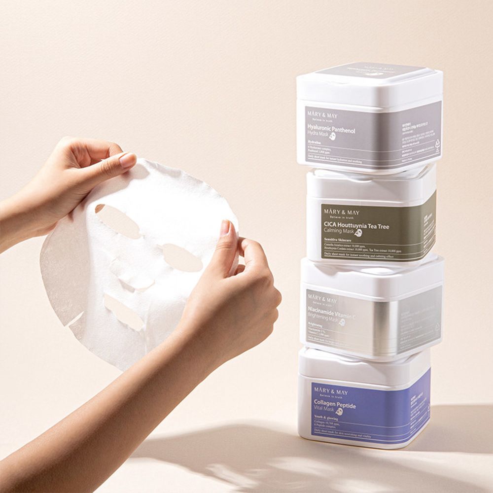 Hände halten eine Gesichtsmaske. Stapel von MARY & MAY Masken-Behältern: Hyaluronic Panthenol, CICA, Niacinamide, Collagen Peptide.