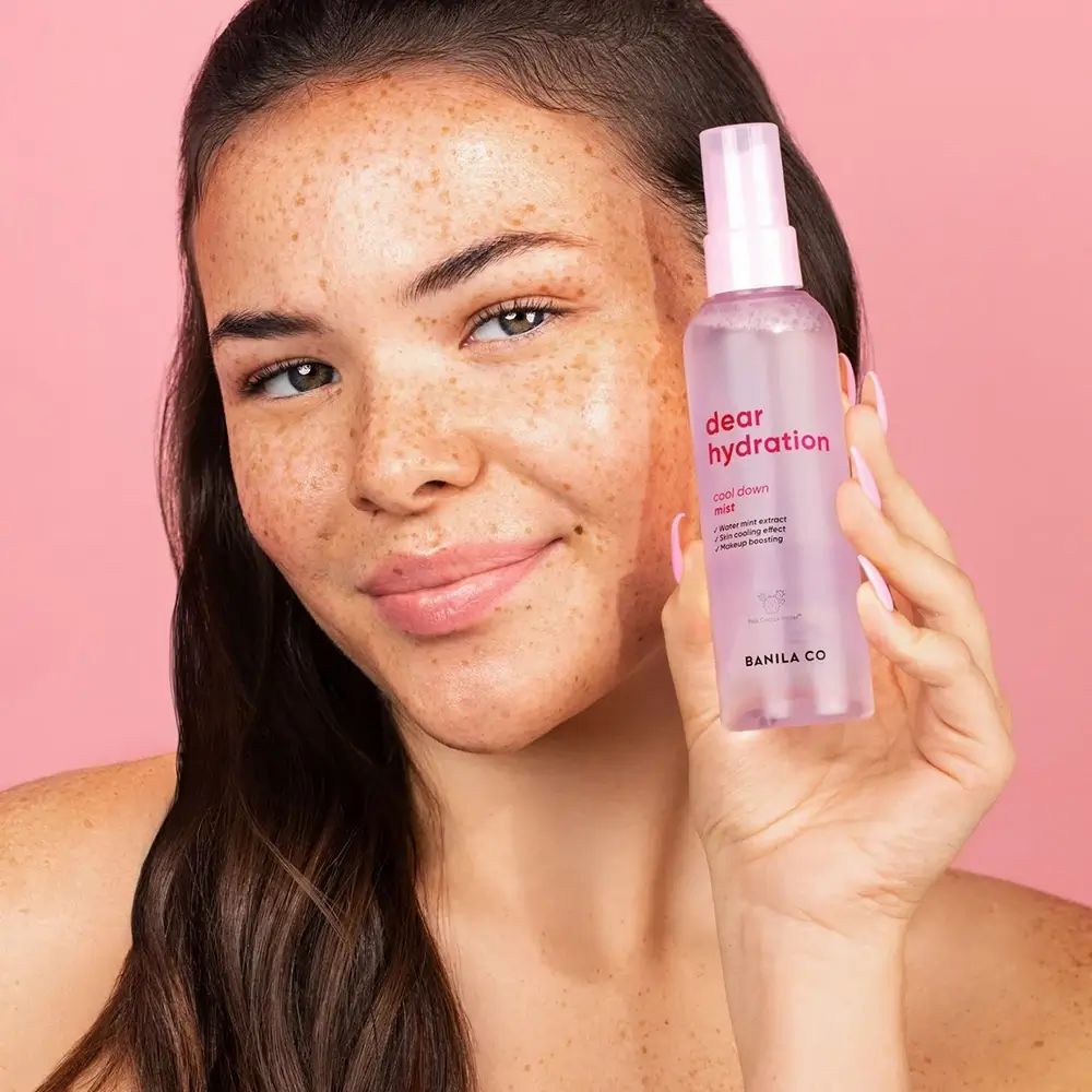 Eine Frau hält eine durchsichtige Sprühflasche mit rosa Flüssigkeit und der Aufschrift „dear hydration cool down mist“ und „BANILA CO“.