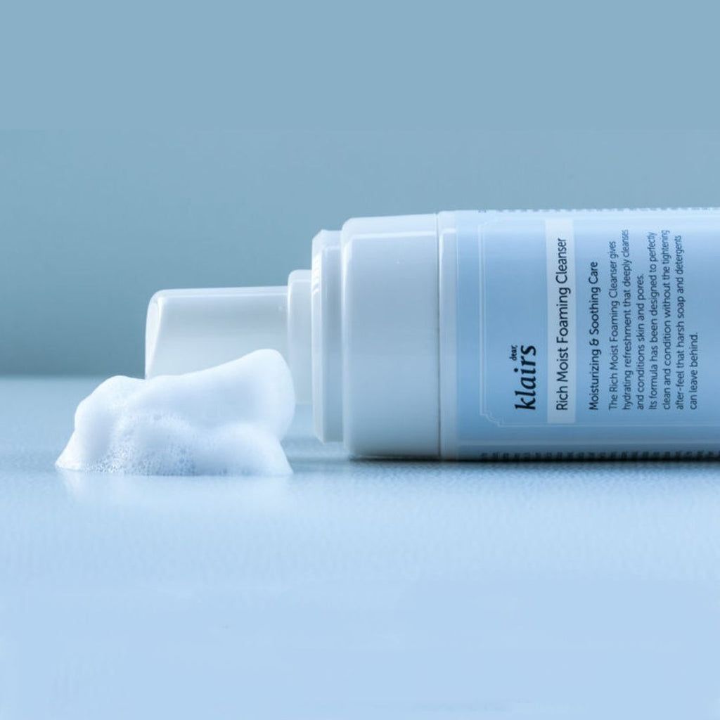 Weiße Flasche Klairs Rich Moist Foaming Cleanser, liegend. Schaum daneben. Blaue Etikett mit Text. Transparenter Deckel.