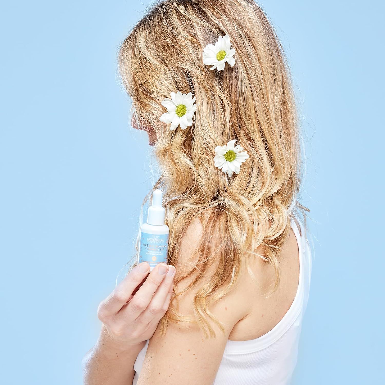 Eine Frau mit blonden Haaren hält einen kleinen Flakon mit Pipette. Blüten sind im Haar.