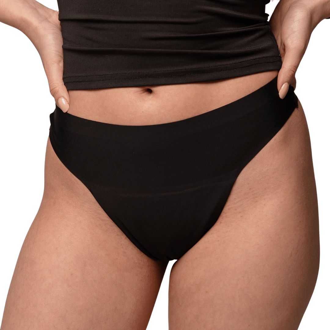 Schwarzer Slip. Das Model trägt ein schwarzes Tanktop. Hände auf den Hüften.