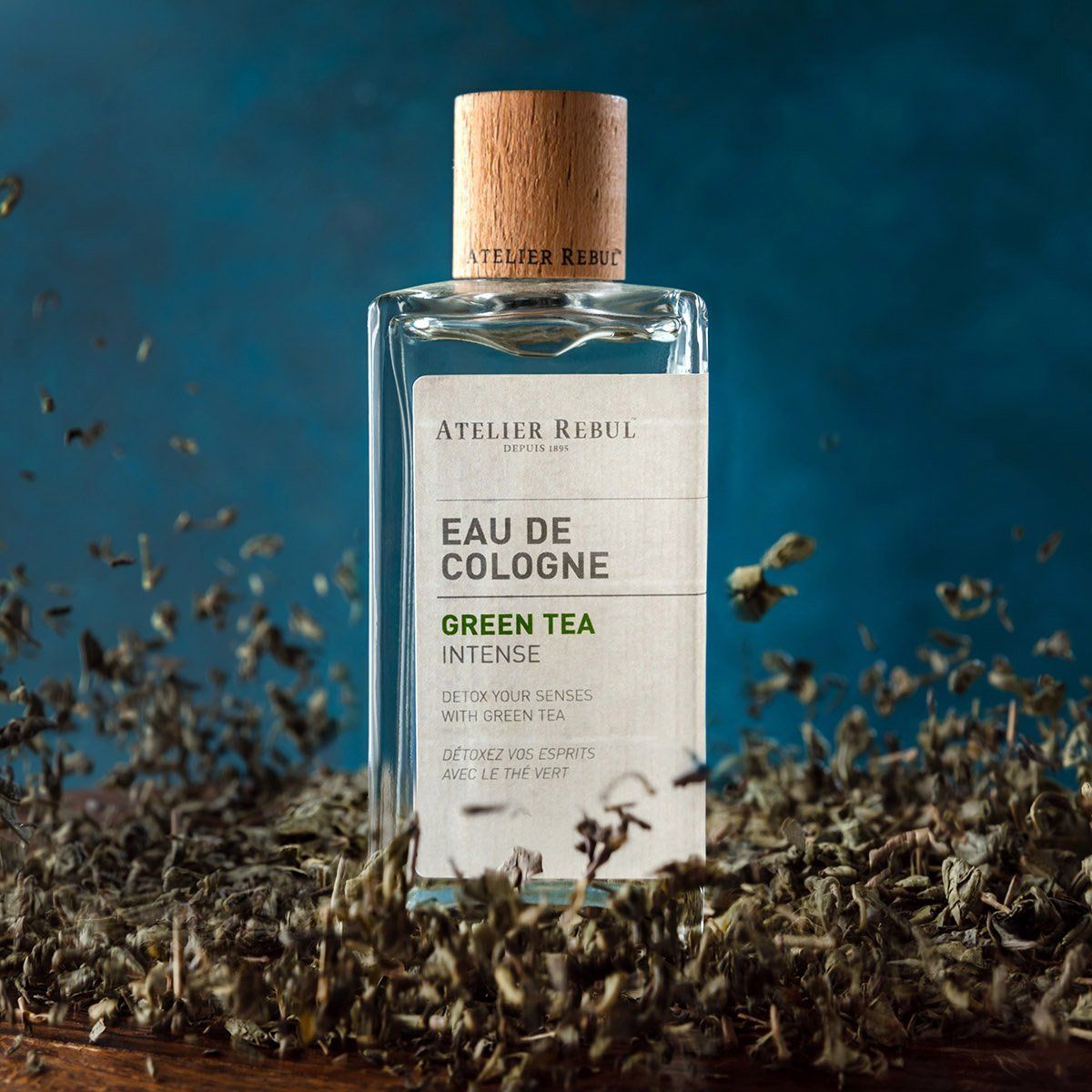 Glasflakon mit Holzverschluss. Aufschrift: Atelier Rebul Eau de Cologne Green Tea Intense. Umgeben von getrockneten Teeblättern.