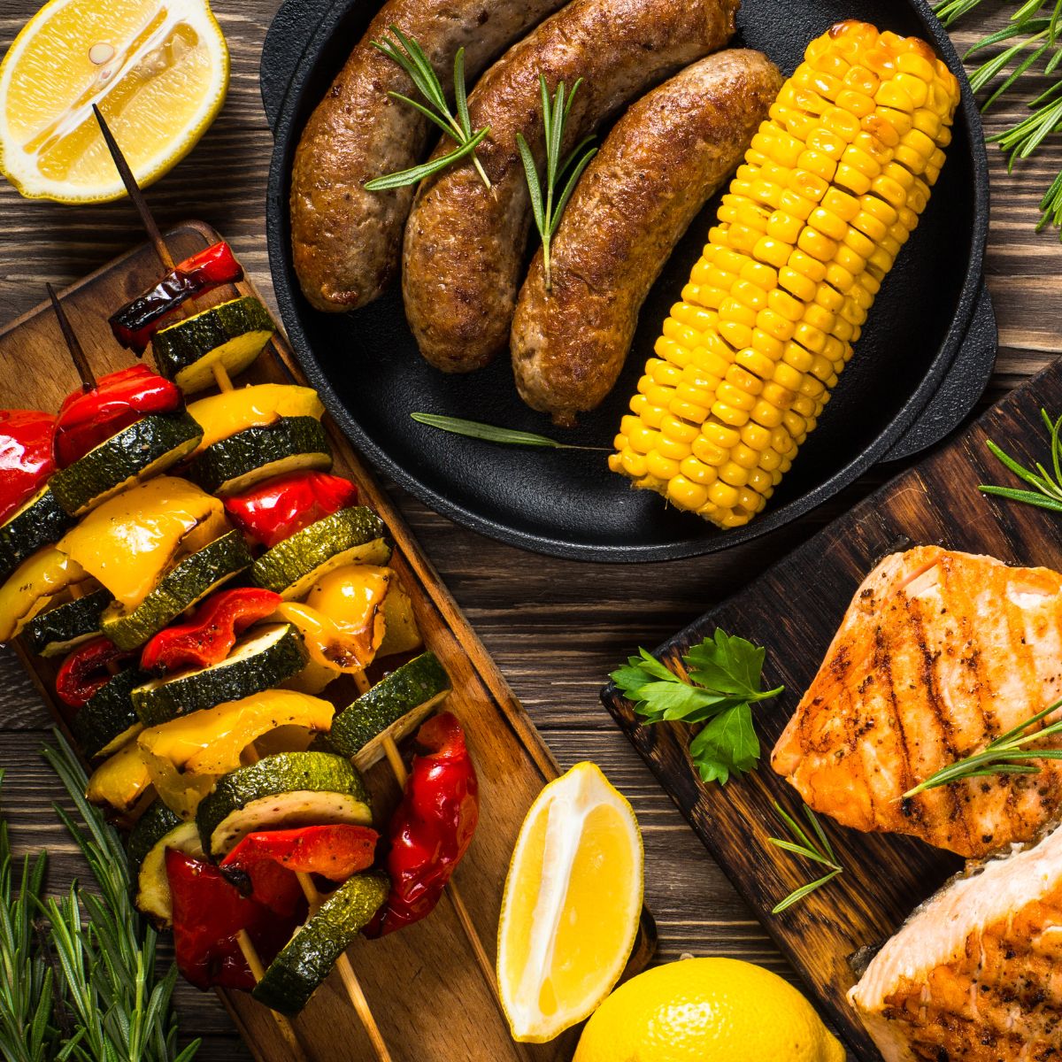 Gegrilltes Essen: Würstchen, Maiskolben, Fisch, Gemüse-Spieße, Zitrone und Rosmarin auf Holzbrettern.