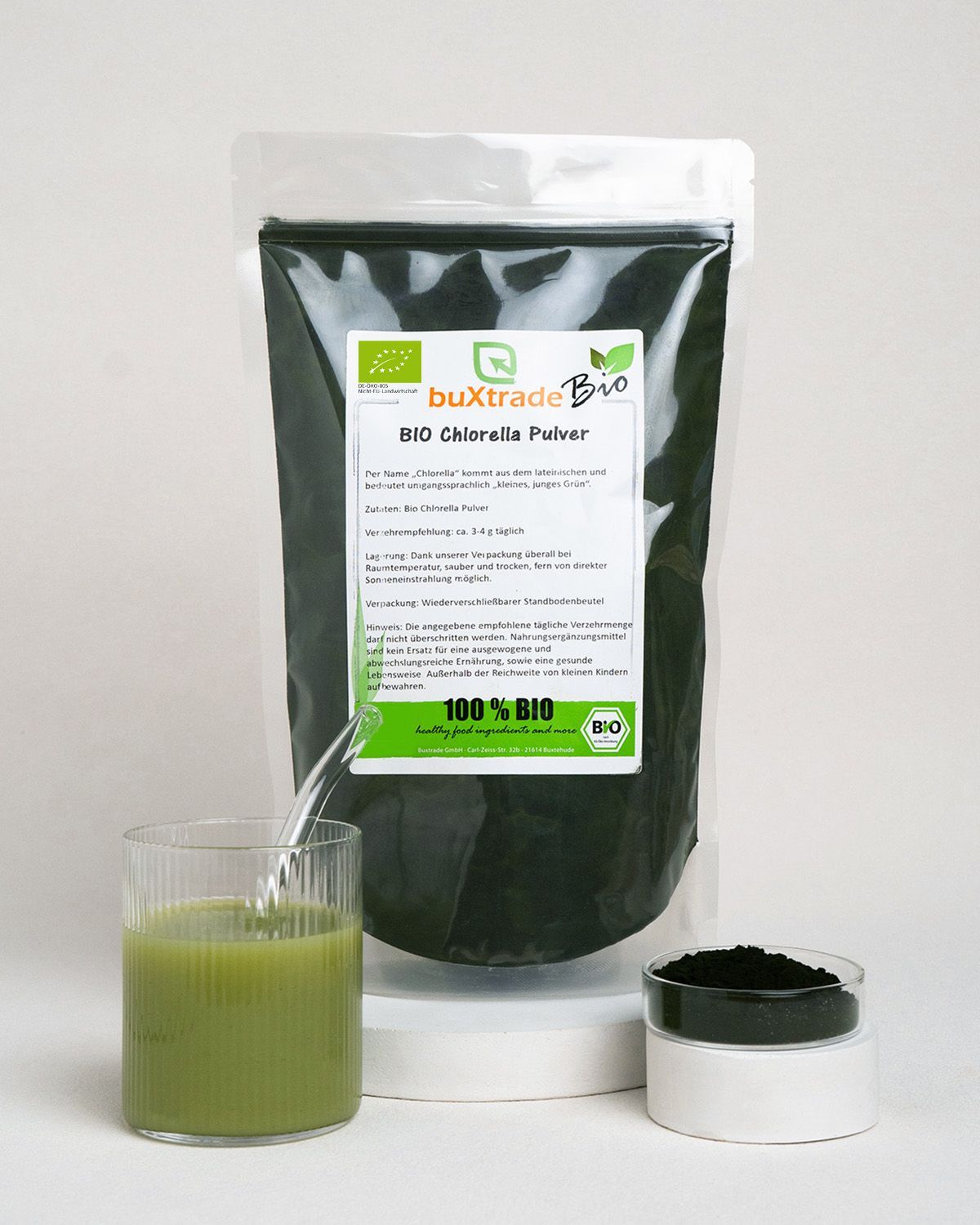 Verpackung von buXtrade Bio Chlorella Pulver neben einem Glas mit grünem Getränk und einer Schale Pulver.
