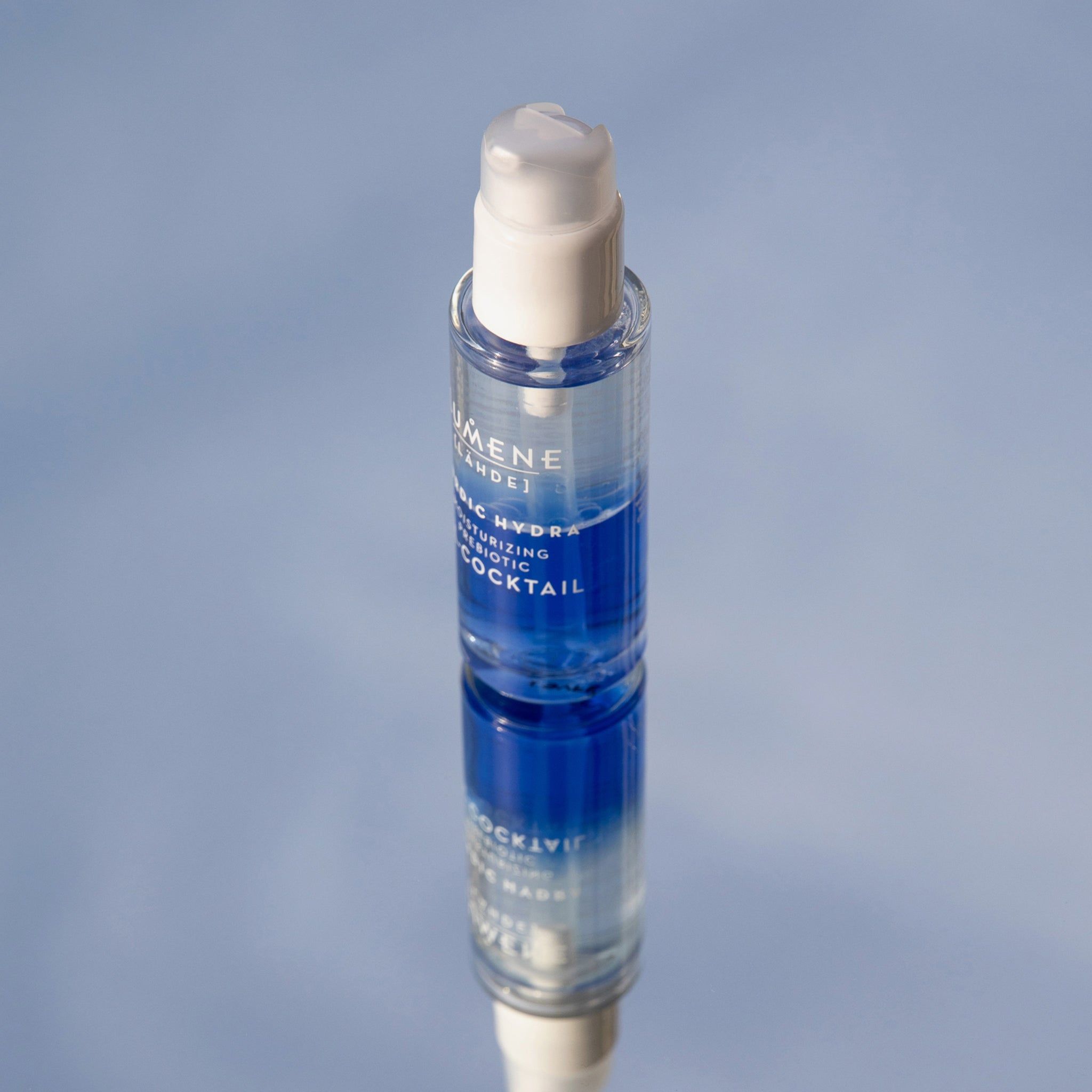 Klarer Flakon mit weißem Pumpkopf, auf einer spiegelnden Oberfläche. Blaue und transparente Flüssigkeit sichtbar. Produktname und Marke.