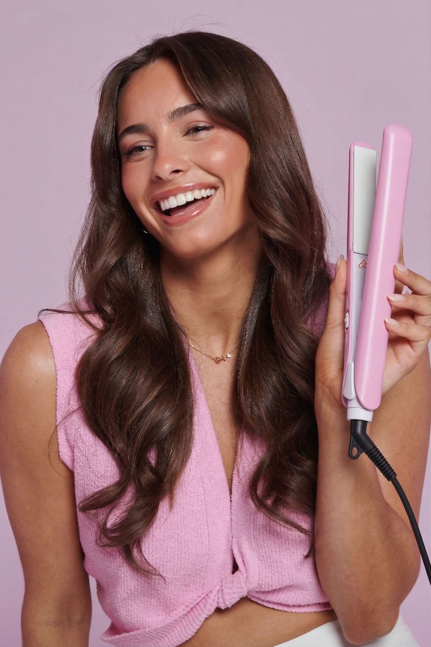 Frau hält rosa Haarglätteisen. Schwarzes Kabel. Frau lächelt. Welliges braunes Haar. Rosa Oberteil.