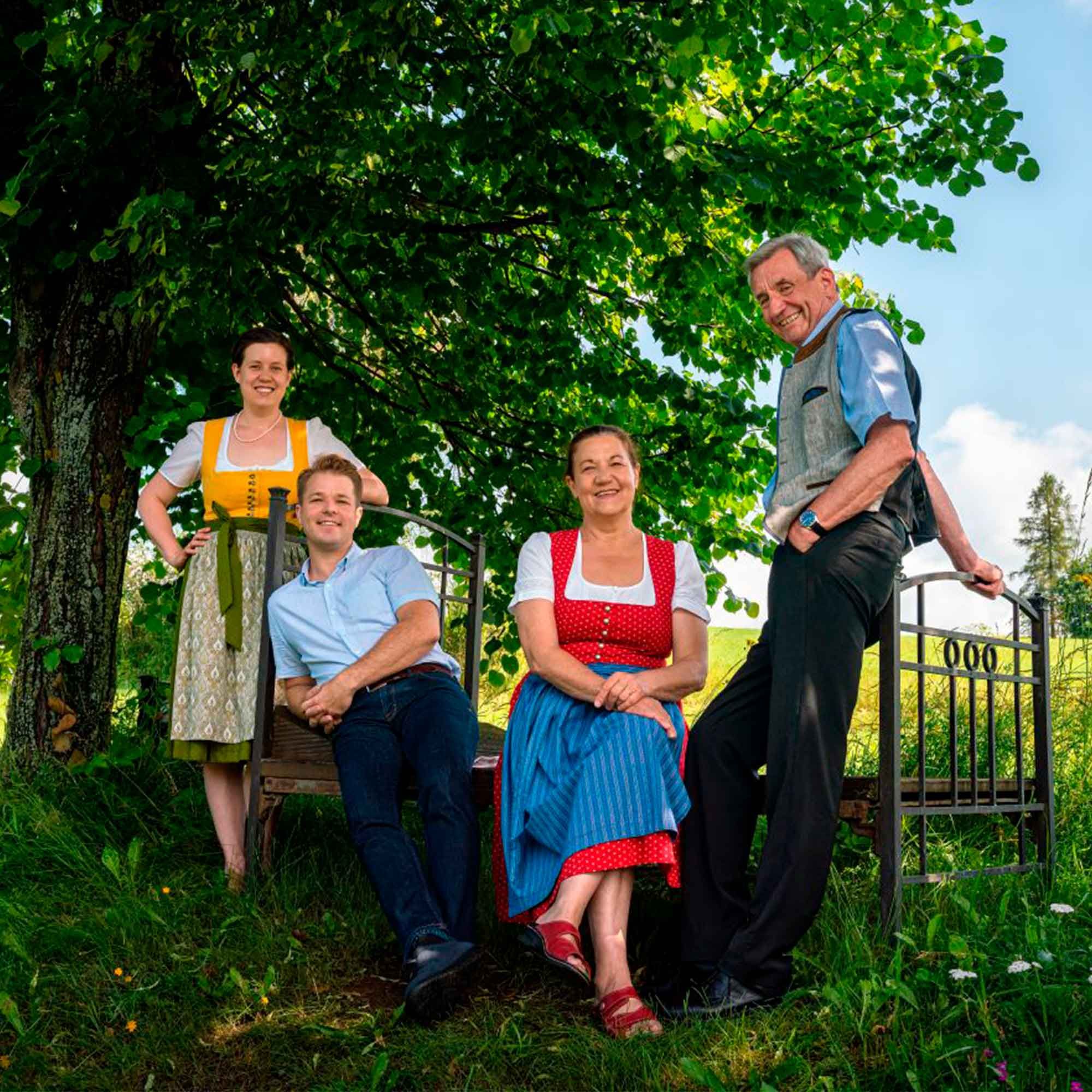 Vier Personen in Tracht sitzen und stehen unter einem Baum auf einer Bank.