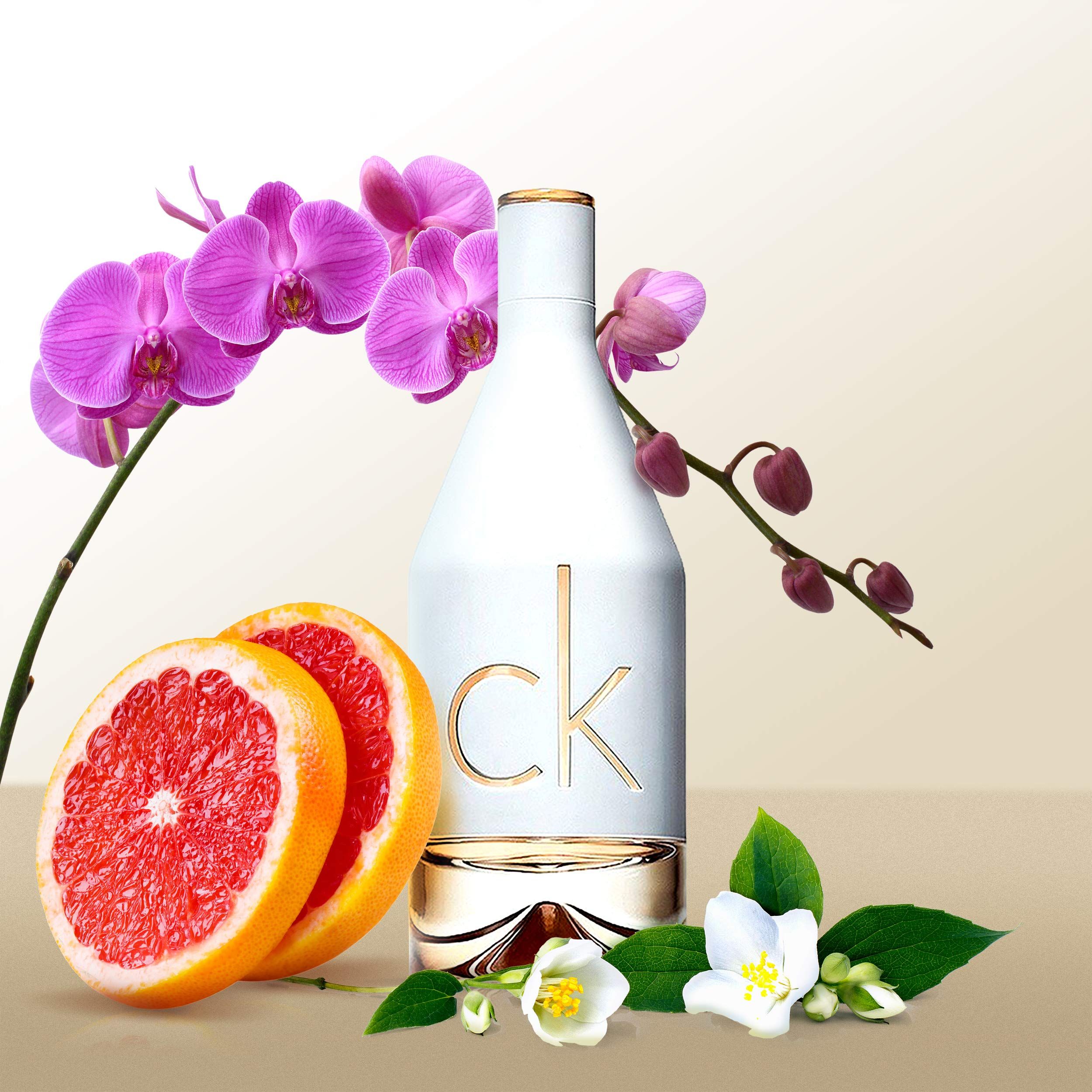 Weiße Parfümflasche mit goldenem "ck"-Logo. Mit Grapefruit, Orchideen und Jasminblüten dekoriert.