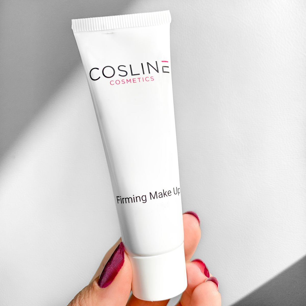 Hand hält eine Tube Cosline Firming Make Up. Hintergrund weiß. Roter Nagellack.