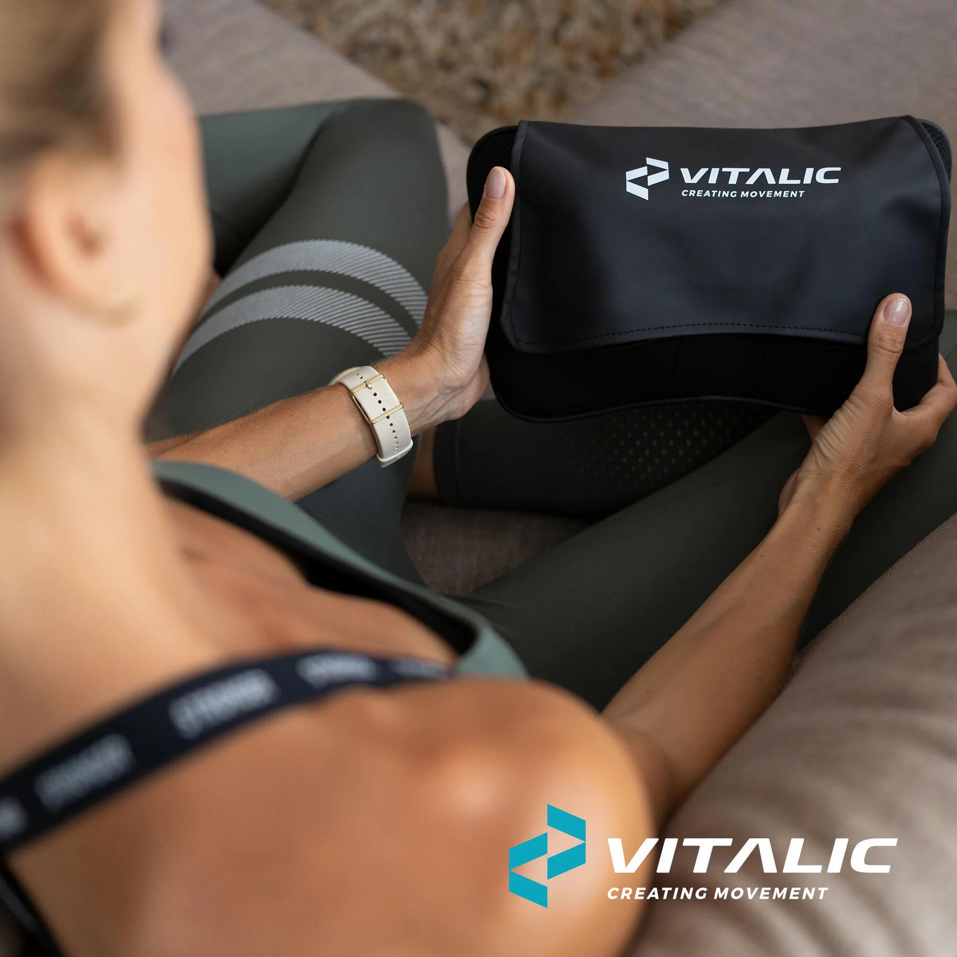 Frau hält schwarzes Massagekissen mit VITALIC-Logo. Kissen mit weißem Logo. Frau sitzt auf Sofa.