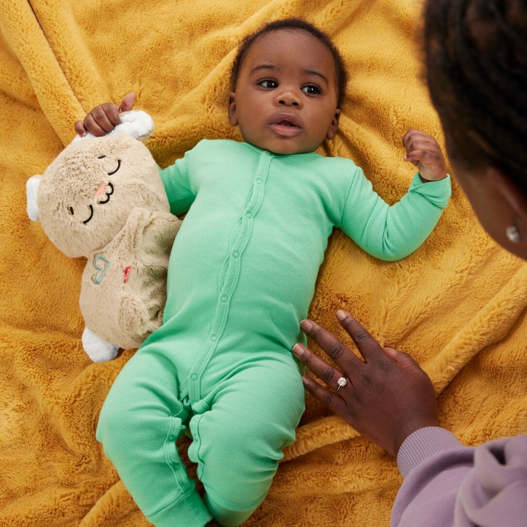 Baby in grünem Strampler hält beiges Schlummer-Häschen. Person berührt das Baby.