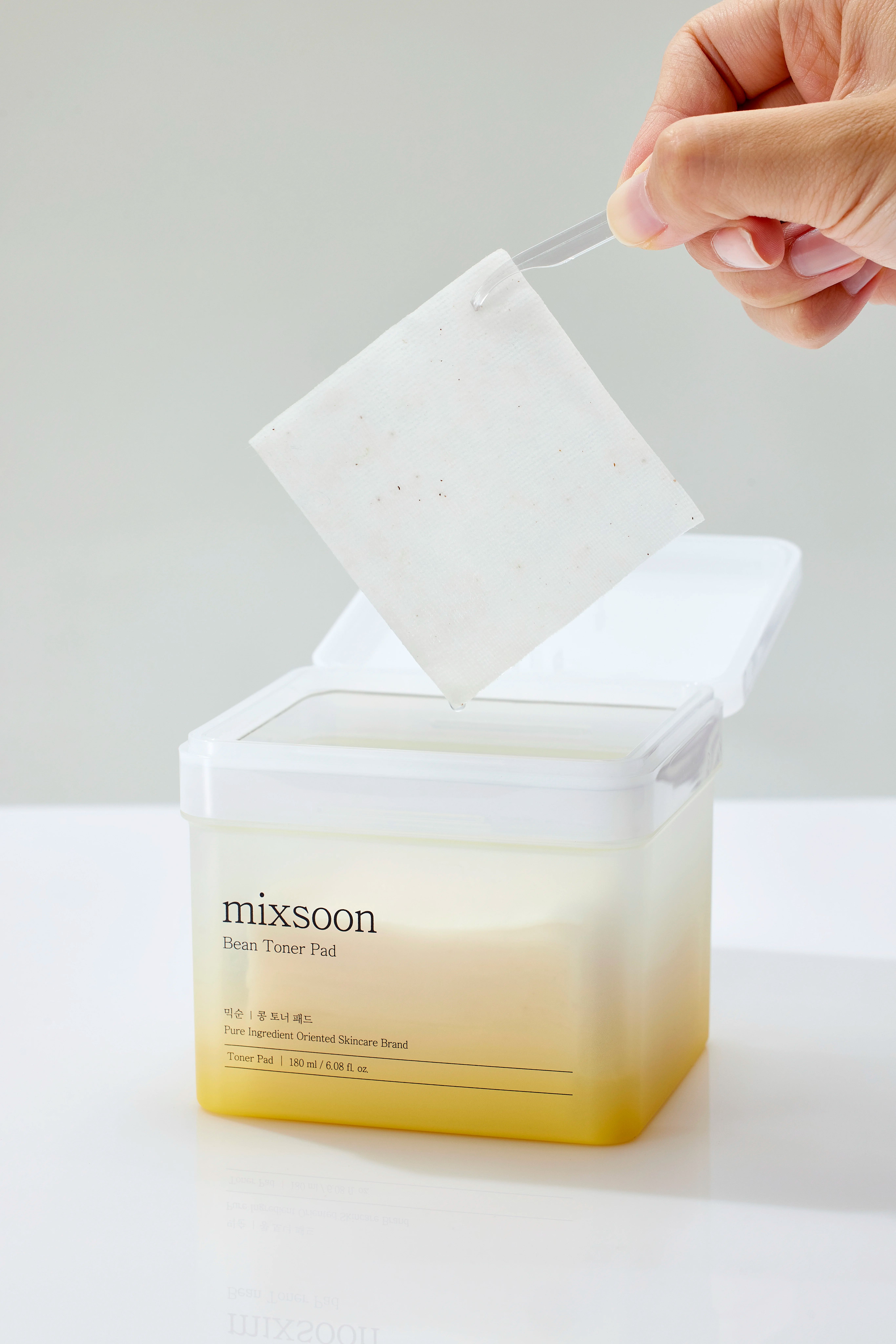 Behälter "mixsoon Bean Toner Pad" mit geöffnetem Deckel. Hand entnimmt ein Pad. Produktname und Details sichtbar.