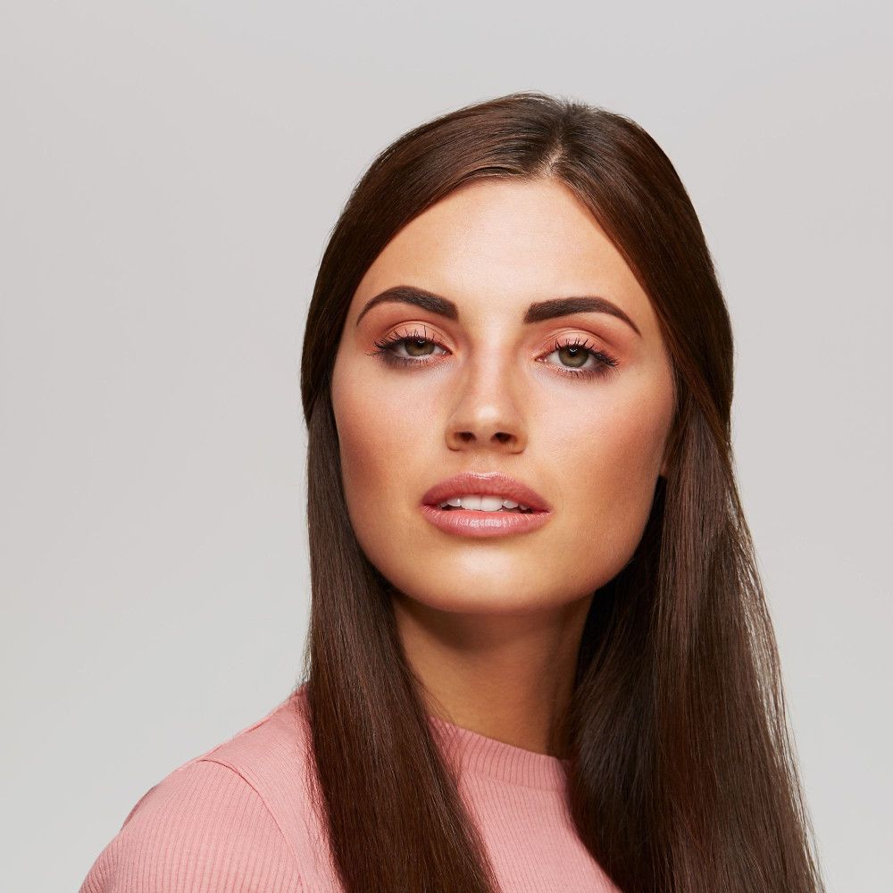 Porträt einer Frau mit langen, braunen Haaren. Sie trägt rosa Kleidung und hat geschminkte Augenbrauen.