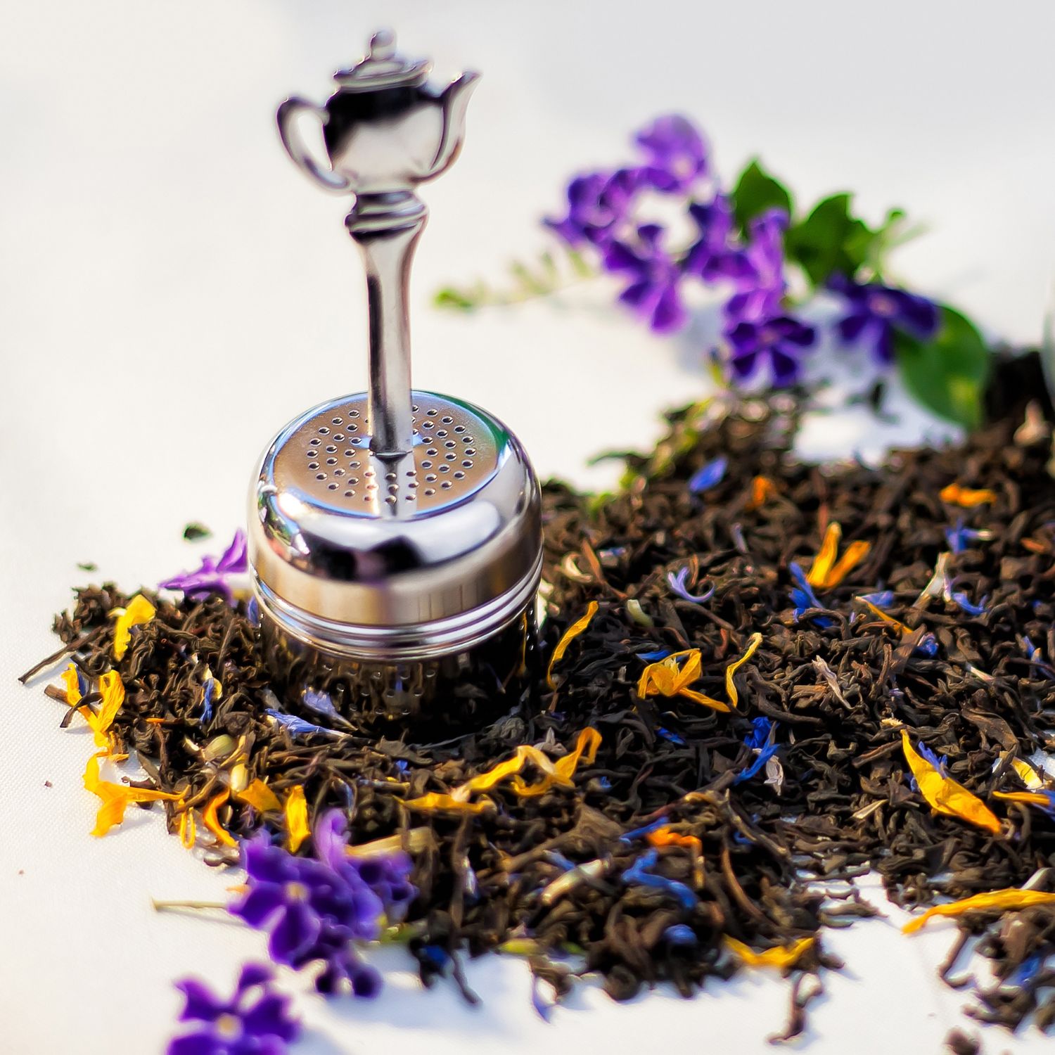 Edelstahl-Teesieb mit Griff. Liegt auf losem Tee mit Blütenblättern. Hintergrund: weiß.